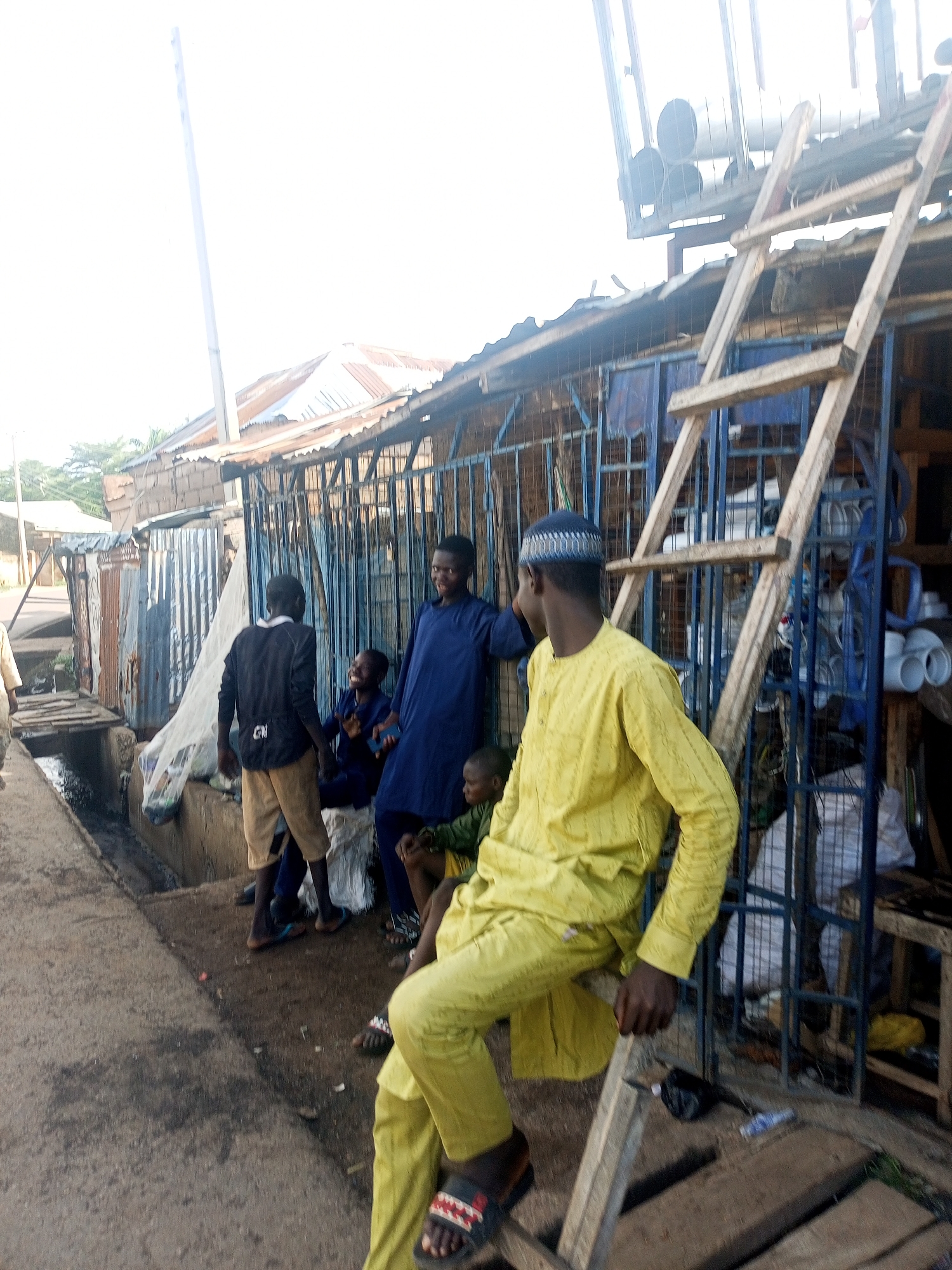 An outdoor scene in an urban or semi-urban environment in Unknown, Unknown, featuring several males and makeshift structures under bright daylight.
In the foreground on the right, a dark-skinned male wearing a yellow long-sleeved tunic, matching trousers, and a blue-patterned kufi cap is seated on a horizontal wooden beam, part of a crude wooden ladder or scaffolding. He wears sandals with red and black straps on his feet. His posture suggests a relaxed lean towards the left.
In the midground, towards the center-left, a group of four males is gathered. One dark-skinned male, wearing a dark blue long-sleeved tunic and trousers, stands smiling, looking towards the right. Another dark-skinned male, wearing a black long-sleeved shirt with a white graphic (possibly "CNI") and light brown shorts, stands facing away from the camera, engaging with the group. A younger dark-skinned male, also in a dark blue tunic, is seated on a white sack, smiling while holding a smartphone. A fourth, younger dark-skinned male, wearing a green top, is partially visible seated on the far right of this group.
The background consists of various informal structures. Immediately behind the midground group is a prominent blue metal grating or gate, possibly for a shop or stall. Behind this grating, white PVC pipes and other dark pipes are visible, suggesting a plumbing or hardware supply business. To the left of the blue grating, several structures made of corrugated metal sheets, some rusted and some weathered, line what appears to be a street. A tall, thin, light-colored pole extends upwards from behind these structures on the far left. Distant corrugated roofs and some sparse vegetation are visible further in the background.
In the foreground on the left, a concrete ditch or open gutter containing dark liquid and debris runs parallel to the ground where the individuals are gathered. The ground itself is a rough, dirty concrete or paved surface. The sky is overexposed due to bright sunlight.