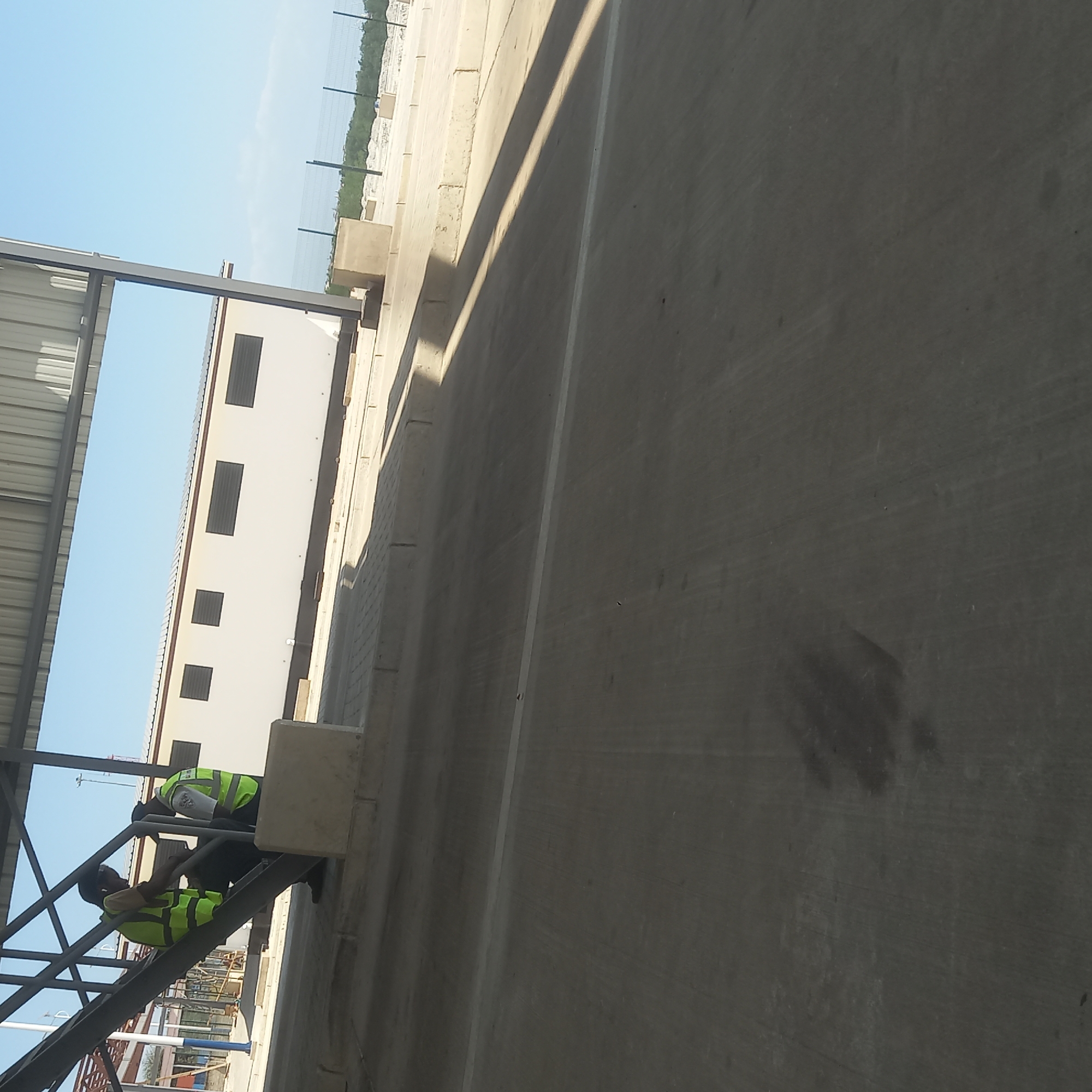 Two individuals wearing high-visibility vests are on a metal staircase. The individual on the left, facing towards the right, is wearing a yellow vest over a tan shirt and dark pants. The individual on the right, facing towards the left, is wearing a yellow vest over a white shirt and dark pants, and appears to be talking to the first individual. They are under a large overhang with a corrugated metal roof. Behind them is a long, white building with dark-tinted windows or louvers. The area in front of the building is paved with concrete and bordered by low concrete curbs. Further in the background, a wire fence encloses an area with vegetation. The scene appears to be in an outdoor, possibly industrial or commercial, setting. The lighting suggests it is daytime. The location in Bububu, Tanzania, is consistent with coastal infrastructure or a port facility.