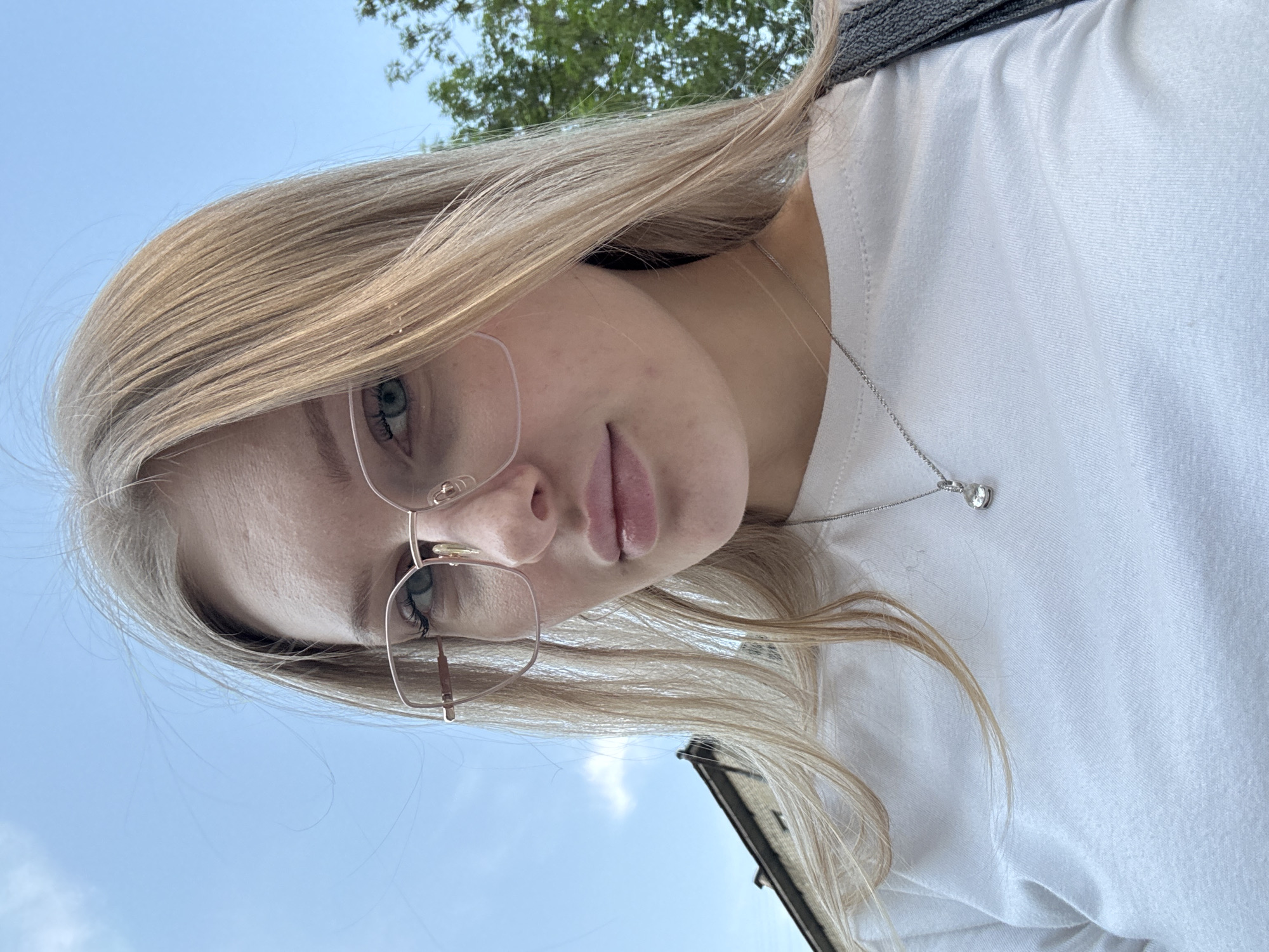 A young woman with blonde hair, wearing glasses and a white shirt, is pictured from a low angle. She is wearing a silver necklace with a pendant. The background shows a blue sky, trees, and a building. The photo appears to be taken outdoors, possibly in an urban setting, but there are not enough clues to determine if this photo was taken in the city of Perm, Russia.