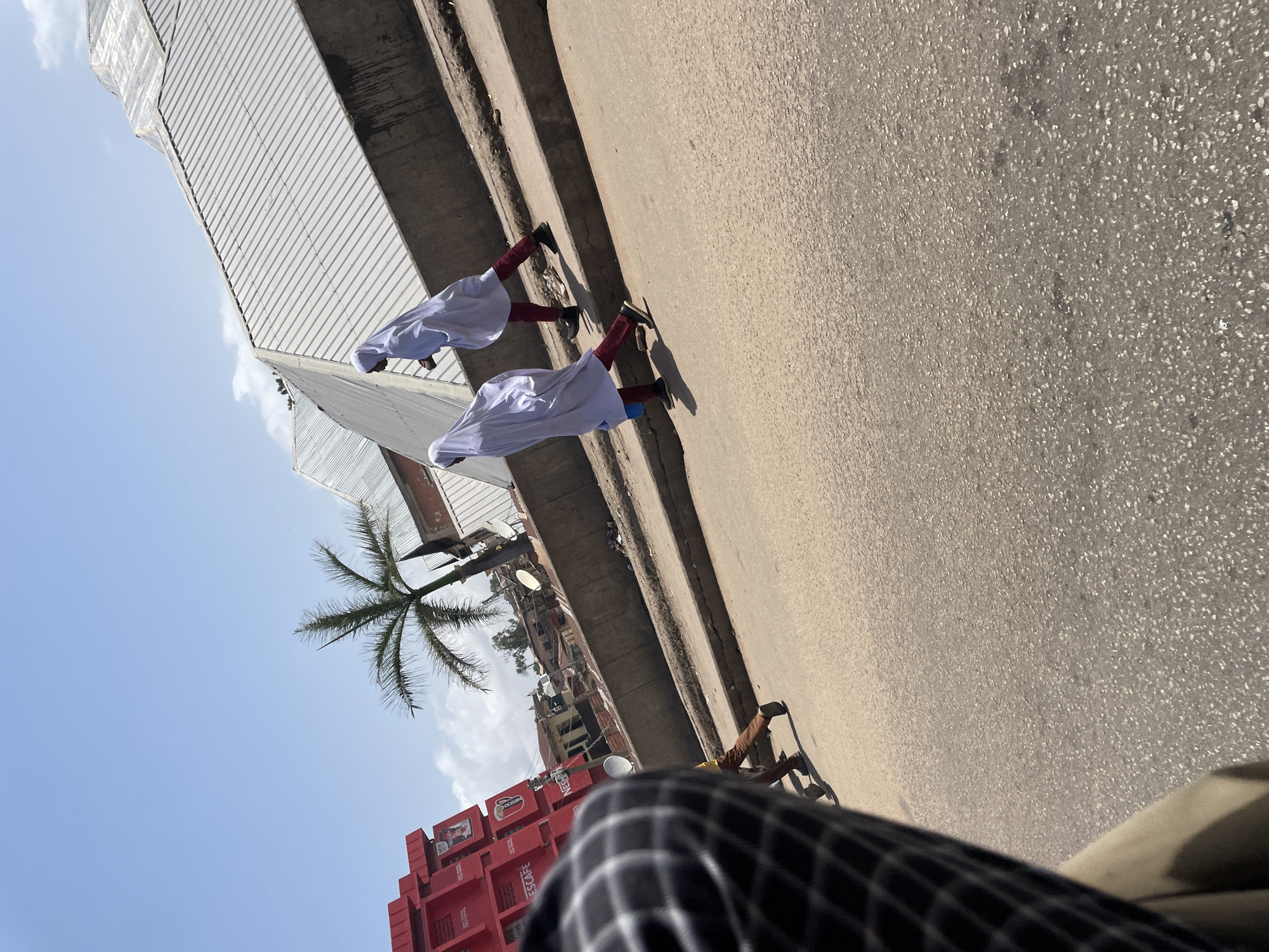 In Jos, Nigeria, on a bright, sunny day, two young women in white hijabs and red trousers walk along a raised sidewalk next to a paved road. They are walking towards the right of the frame. In the background, a tall palm tree stands next to a building with a corrugated metal roof. To the left, a red building with signs is visible. The foreground shows a partial view of a person's leg and clothing, suggesting the photo was taken from a moving vehicle.