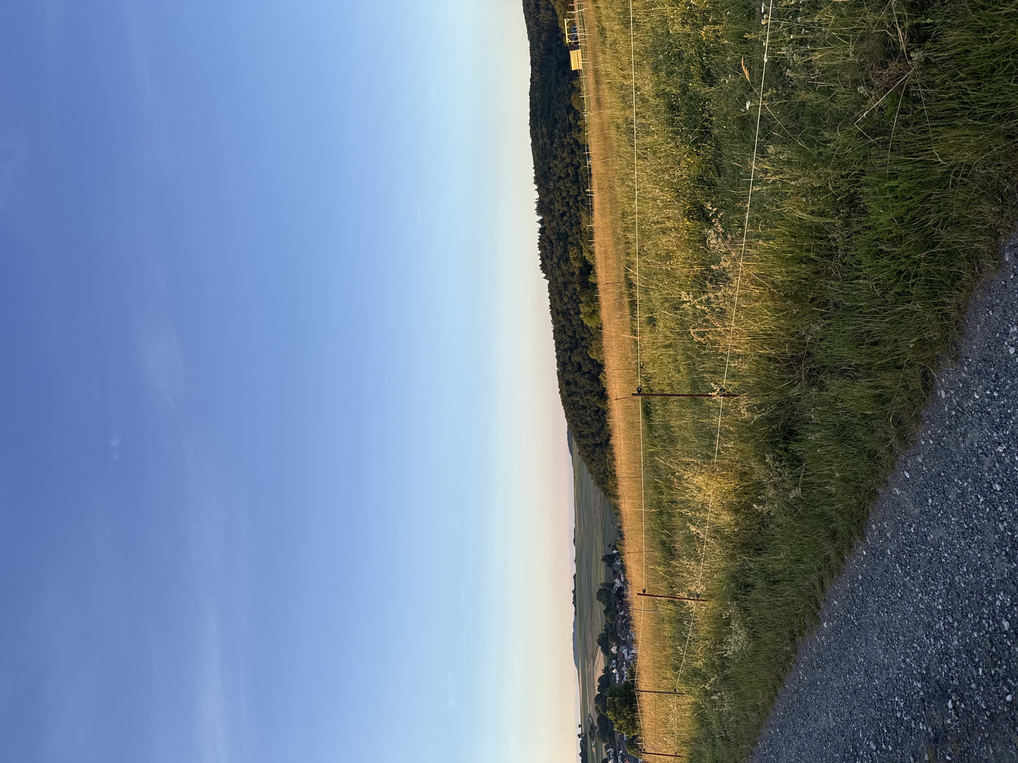This image depicts a rural landscape with a clear blue sky at dusk. In the foreground, a gravel path runs along the left side, bordering a field of tall, golden-brown grass. A simple wire fence with wooden posts runs through the field, separating it from the path. In the mid-ground, a small village with red-tiled roofs is nestled among rolling hills, with patches of green fields and trees. A dense, dark green forest covers a prominent hill on the right side of the image. The light suggests the sun is low on the horizon, casting a warm glow on the landscape. The scene is serene and depicts the outskirts of the city of Oberquembach, Germany.