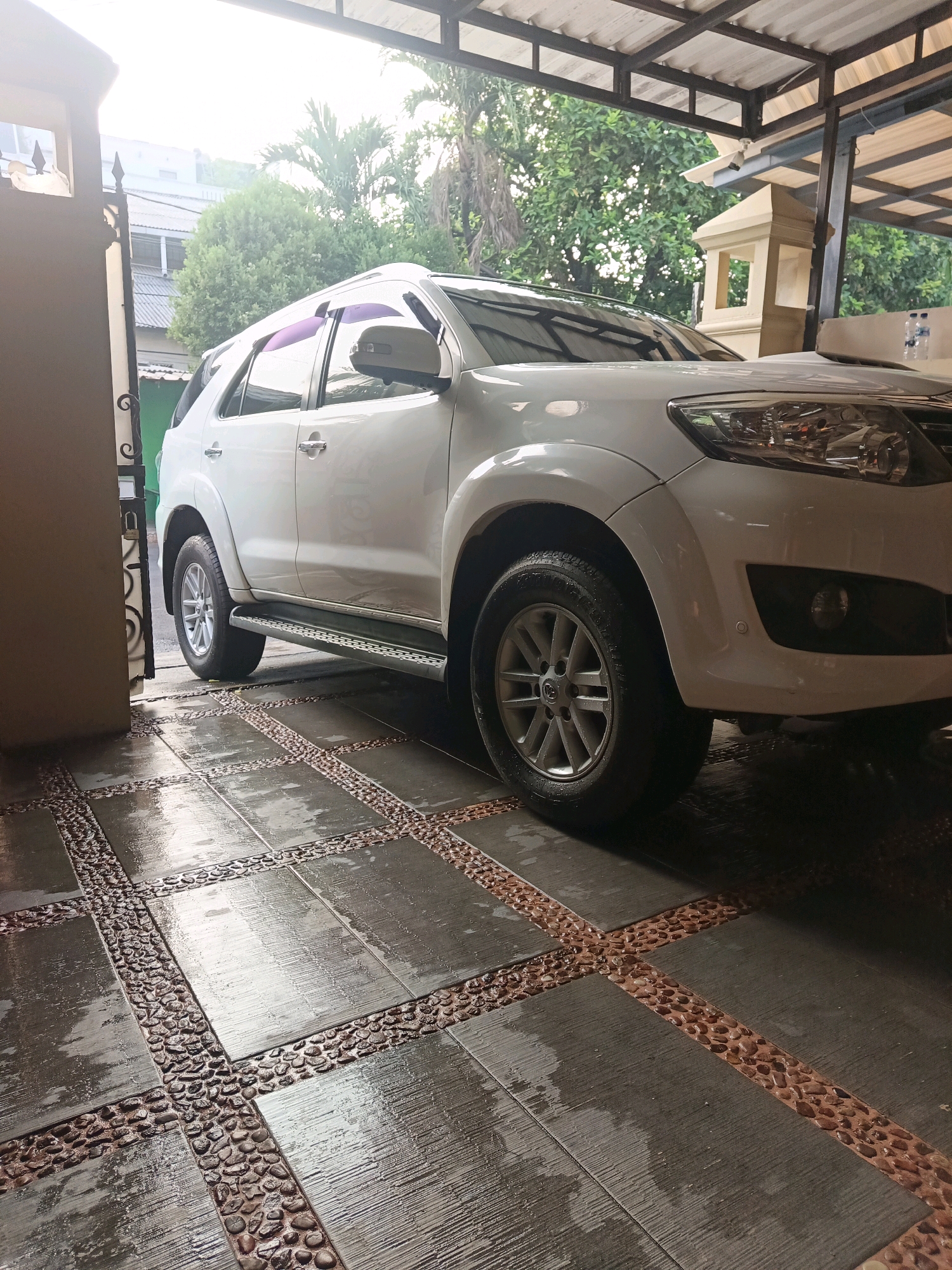 A white SUV is parked on a wet, patterned stone driveway under a covered patio in North Jakarta, Indonesia. The vehicle is facing to the right, with its front left wheel visible. The patio roof is constructed of metal beams and translucent panels, allowing natural light to filter through. Lush green trees and foliage surround the patio, with a glimpse of a residential building in the background. The driveway is paved with dark, rectangular tiles interspersed with bands of small, rounded pebbles, all glistening with moisture. A partial view of an ornate gate is visible on the left side of the frame.