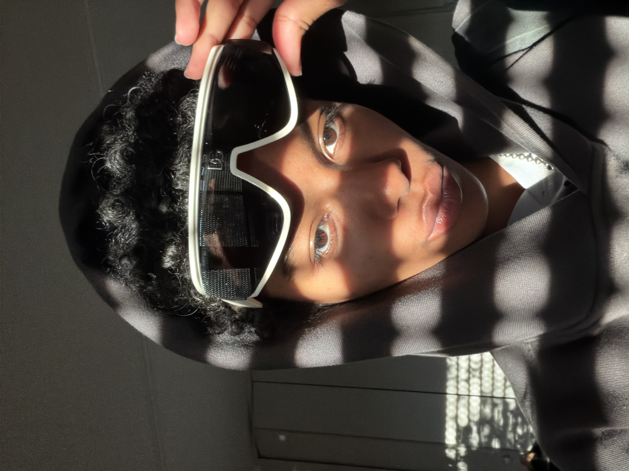The image depicts a person wearing a hooded sweatshirt and oversized white sunglasses, with the sunglasses being held slightly above their eyes by one hand. They have dark curly hair and brown eyes, and their face is partially obscured by shadows. The background includes a wall with horizontal panels. The image appears to have been taken indoors. There is no information in the image that indicates the location is Champs-sur-Marne, France.
