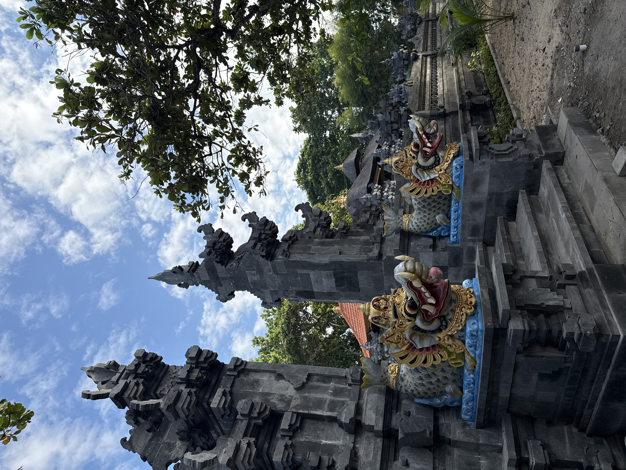 The image depicts a detailed view of a Balinese Hindu temple, likely situated in Kuta Selatan, Indonesia. The scene is dominated by dark grey stone structures characteristic of Balinese temple architecture, including elaborate tiered gates and walls. In the foreground, two vibrant, mythical guardian figures are prominently featured. These statues resemble dragon-fish creatures, with intricately carved golden crowns, red open mouths revealing teeth and a pink tongue, and scale-like bodies painted in grey, gold, and blue, resting on bright blue pedestals.
The structures themselves are composed of precisely cut and stacked stone blocks, forming complex patterns and decorative elements that ascend skyward, culminating in spires. To the left, a tall, multi-tiered structure partially frames the view. To the right, another similar, slightly less prominent structure is visible. Stone steps lead up to the platforms where the guardian statues are positioned.
The ground in the immediate foreground and to the right consists of light brown earth and scattered small rocks, with some sparse green foliage and a small palm-like plant suggesting ongoing or recent landscaping. In the midground and background, dense green trees with varied foliage create a natural backdrop. Visible through and above the stone structures are parts of additional temple buildings, including a structure with a reddish-orange tiled roof and others with traditional dark thatched roofs, all surrounded by abundant greenery. The sky above is a clear blue, dotted with scattered white cumulus clouds. No people are visible in the scene.