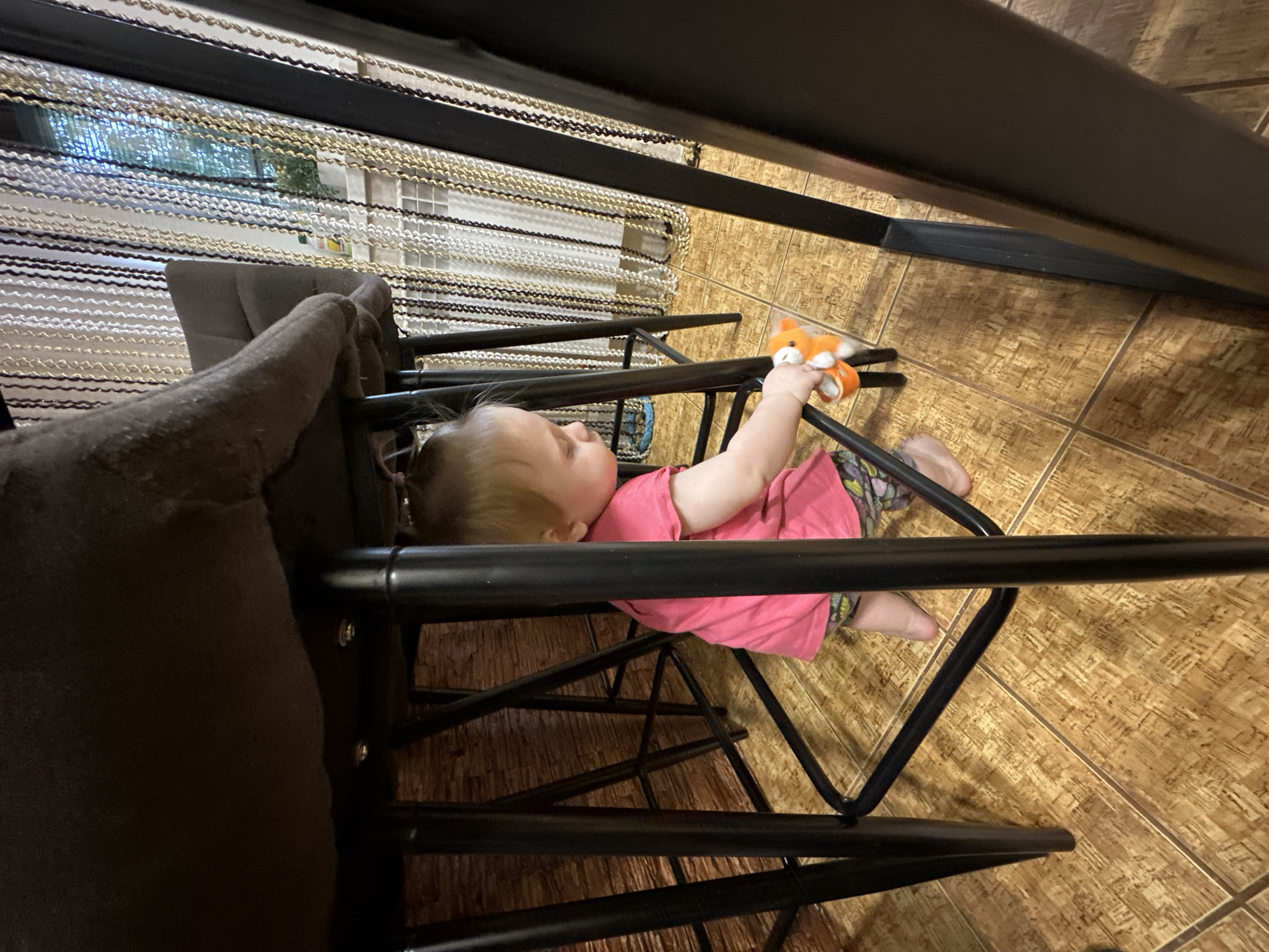 A young child, possibly a toddler, is lying on their side on a black metal chair. They are wearing a pink shirt and patterned leggings. The child is holding a small orange stuffed fox toy in their right hand. Their head is resting on their left arm. The chair appears to be part of a set of furniture. In the background, a window is visible with beaded curtains in front of it. The flooring is tiled and appears to be made of large, square tiles in a warm, earthy tone. The overall lighting suggests it might be daytime indoors. The scene is likely a home environment in Sayanogorsk, Russia.