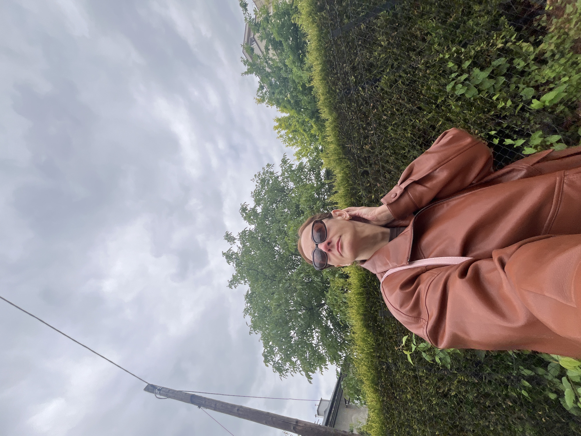 A woman with brown hair, wearing sunglasses and a brown leather jacket, stands in front of a hedgerow with a wire fence. She is touching her ear and looking upwards. A utility pole with a wire stretches across the cloudy sky. Behind the hedgerow, trees and a building are visible. The scene is outdoors in an urban environment.