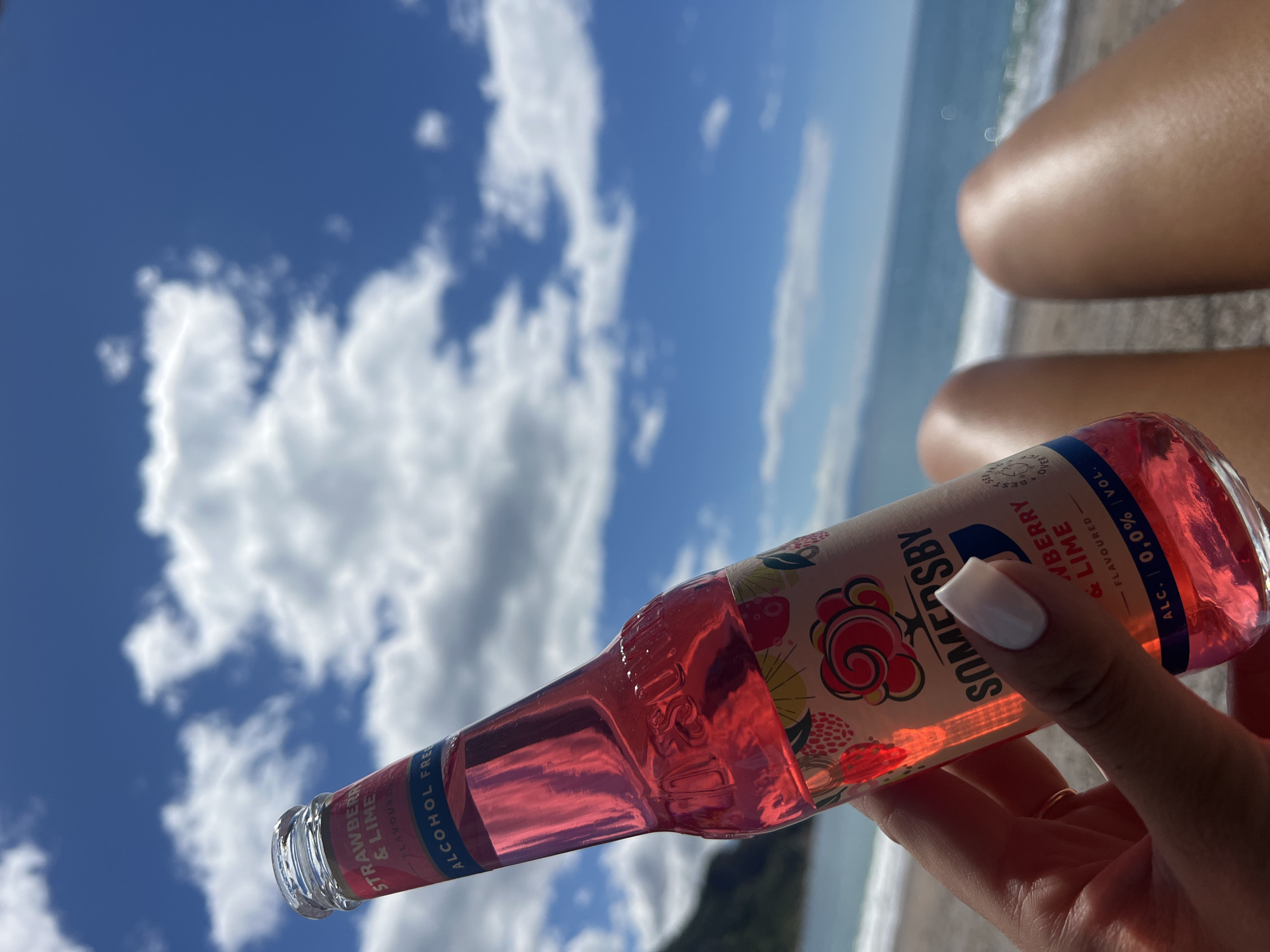 A person is holding a bottle of Somersby Strawberry & Lime flavored non-alcoholic beverage in front of a beach landscape. The drink bottle is pink and white, with text stating "STRAWBERRY & LIME" and "ALCOHOL FREE". The person's hand is visible, with a white fingernail. Behind the bottle, a beach scene unfolds with a turquoise ocean, a sandy beach, and a blue sky with white, fluffy clouds. Part of the person's legs are visible in the upper right portion of the frame. The location is Prijevor, Montenegro.