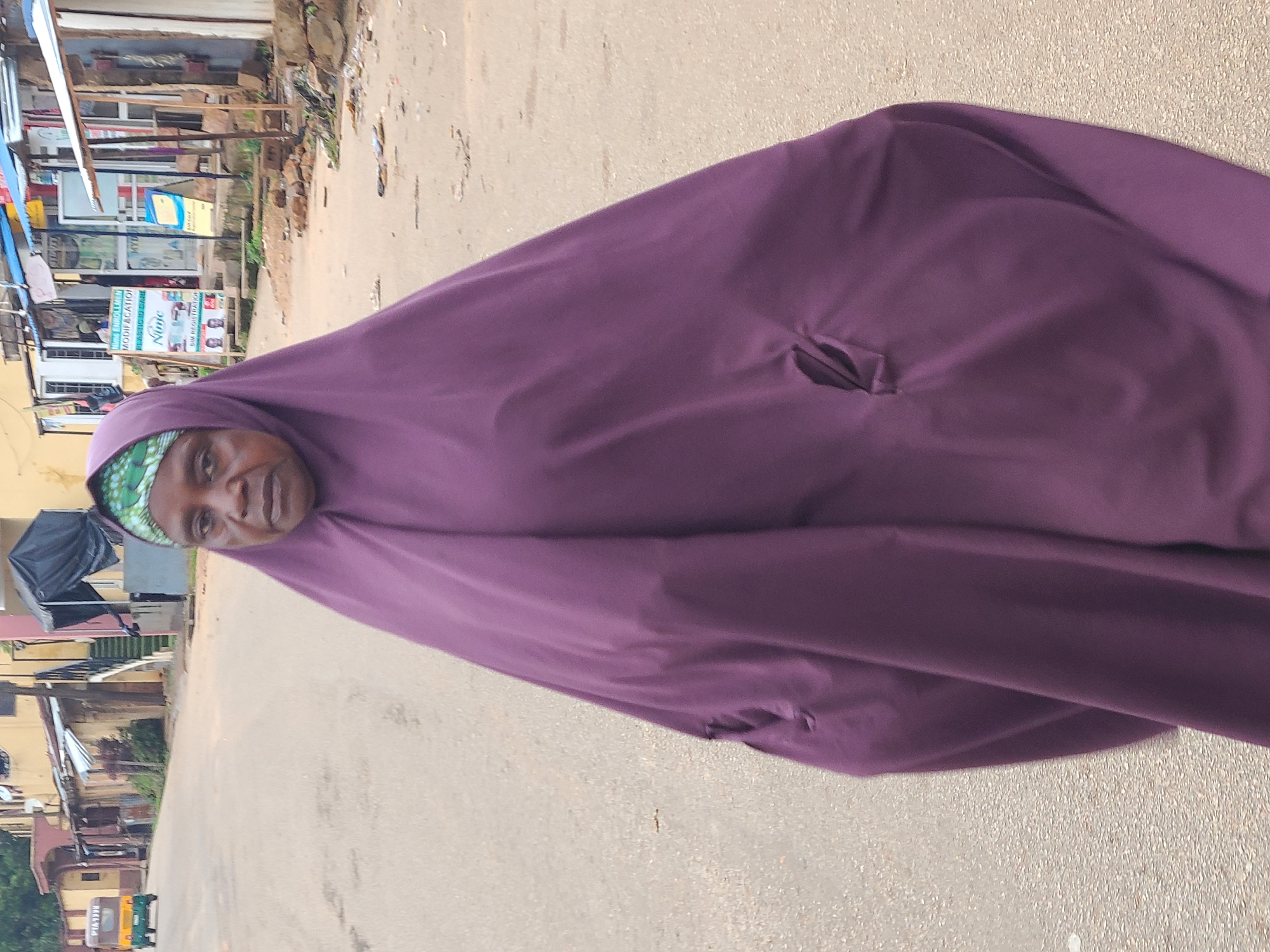 A woman wearing a purple hijab and dress stands on a paved street in Jos, Nigeria. The woman's face is visible, and she is looking towards the camera. The street is lined with buildings and shops on either side. In the background, a yellow and green auto-rickshaw is visible, as well as people walking and standing near the shops. The overall scene depicts a typical street view in a Nigerian city.