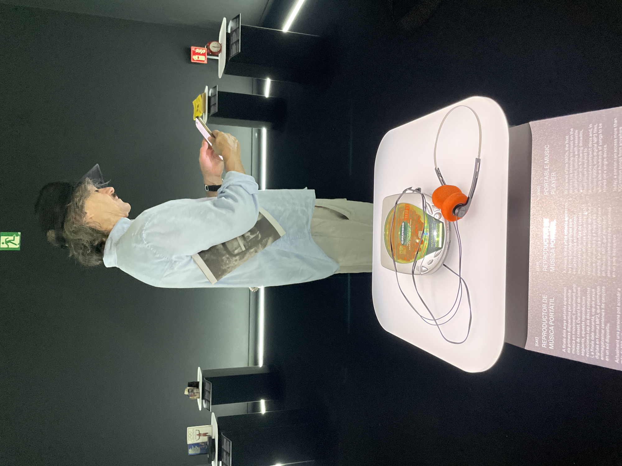 The image depicts an indoor exhibition setting, likely a museum, with a minimalist aesthetic. In the foreground, a white, illuminated pedestal displays a portable music player with orange headphones. The player appears to be a CD player, with a visible disc. To the right of the player, text in Catalan and English describes portable music players. In the mid-ground, a person wearing a light blue shirt, khaki pants, and a black hat is looking down at a smartphone they are holding. They are also holding a booklet or pamphlet. The background is dark, with illuminated displays featuring various objects on black pedestals. One display shows what appear to be small books or boxes, and another features a red alarm clock. The scene is dimly lit, with accent lighting highlighting the exhibits and the floor. The presence of Catalan text suggests the location is in Catalonia, Spain, and the overall presentation is consistent with an exhibition in Barcelona, possibly related to design, technology, or cultural history.