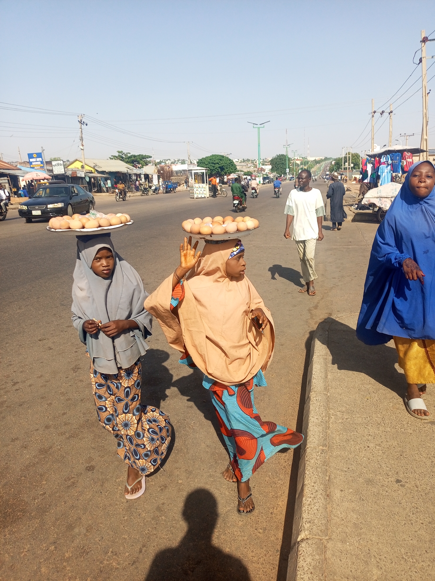 This photograph captures a bustling street scene in Tempure, Nigeria, under a clear, bright sky, indicative of mid-morning or early afternoon due to the distinct shadows cast by people and objects.
In the foreground, two young women are prominently featured, each balancing a large, flat tray full of light-colored eggs on their heads, a traditional method of street vending. The woman on the far left wears a grey hijab and a patterned skirt in browns and blues, while the woman in the center has an orange-beige hijab and a vibrant blue and orange patterned skirt, with one hand slightly raised to steady her load. To their right, another woman, dressed in a blue hijab and yellow trousers, walks along the concrete curb, looking towards the camera.
The background reveals a lively street lined with various market stalls and buildings. Several men are visible; some walk along the road, while others ride motorcycles, contributing to the busy atmosphere. A black sedan is seen on the paved road, which appears to be a main thoroughfare. Overhead, numerous power lines stretch across the sky, supported by utility poles. Streetlights are also visible further down the road.
Notable details include a white sign displaying the text "NASFAT" and a partially visible blue and white sign that appears to read "BUDGET WITH ANB..." or similar. The scene depicts daily life and commercial activity in an urban or semi-urban setting, highlighting the everyday routines and the common practice of carrying goods on one's head for sale. The overall impression is one of movement and typical market day commerce.