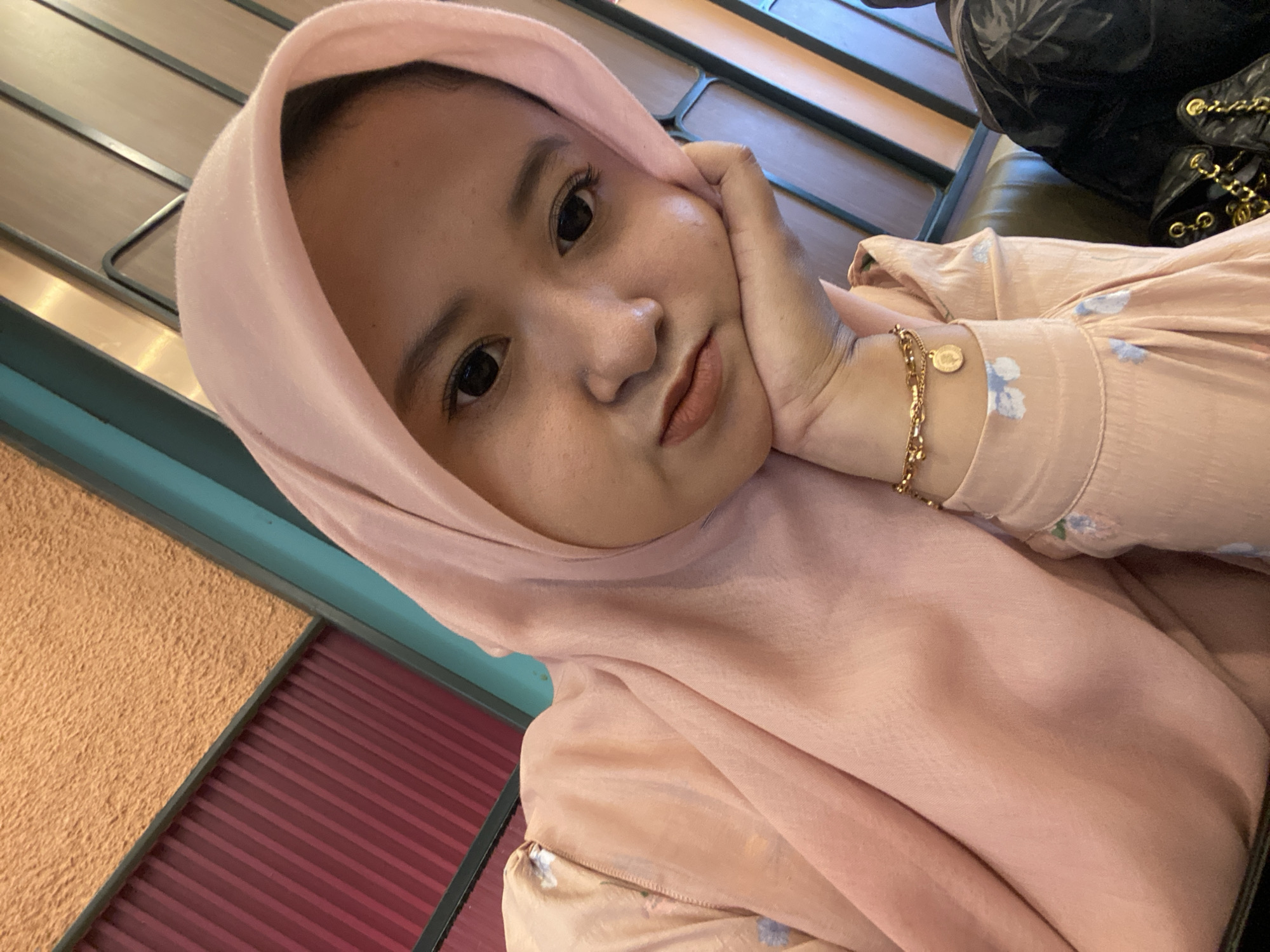 A medium-shot, indoor, eye-level selfie captures a young woman wearing a light pink hijab and a matching pink dress with subtle floral patterns. She is resting her chin on her hand, with her left arm bent and resting on her chest. A gold bracelet adorns her left wrist. The background consists of a wall with textured orange panels and vertical red elements, intersected by teal-colored trim. The lighting is warm and appears to be artificial. The image is undated and provides no explicit location information, but its style suggests a casual setting within the city of Sidoarjo, Indonesia.