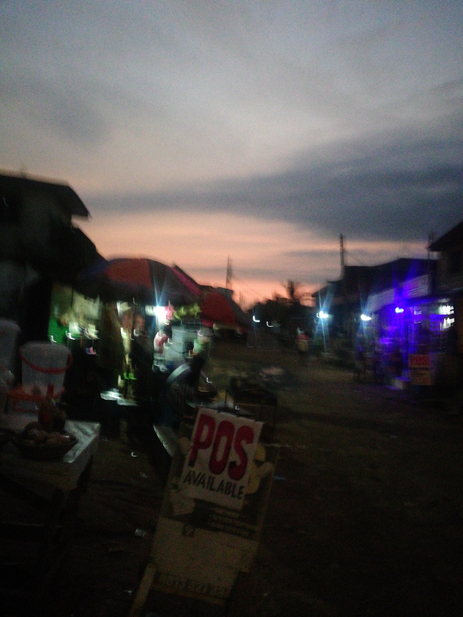 The image shows a blurred, dark street scene at dusk in an urban environment. The sky is a mix of orange and gray, suggesting either sunset or sunrise. In the foreground, there is a sign that reads "POS AVAILABLE". Behind the sign, there are street vendors operating under tents, illuminated by artificial light. Further down the street, buildings and streetlights are visible, with some storefronts displaying blue neon lighting. Overall, the scene appears to be a street market or commercial area at twilight in the city of Unknown, Unknown.