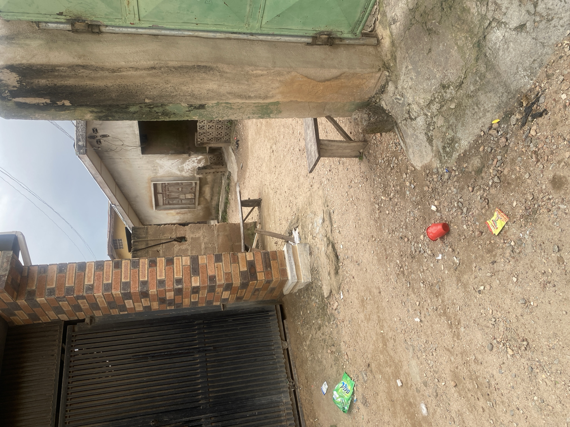 This image depicts a dusty, outdoor scene in Abeokuta, Nigeria, with elements suggesting a somewhat neglected residential area. In the foreground, a dirt and gravel ground is littered with debris, including a crumpled green plastic bag, a red plastic cup, and a yellow and red packaged snack. A section of a rough, unpaved concrete surface is visible on the right, stained with moss and dirt.
Dominating the left side is a tall, brick pillar with alternating red and dark brown bricks, topped with a concrete cap. Adjacent to this pillar is a tall, black metal gate. To the right of the pillar, a portion of a weathered, green metal door is visible, featuring vertical bars and a prominent handle.
Further into the scene, a light-colored building with a sloped roof is partially visible. It has a single window with wooden shutters. Another, more weathered building with a patterned concrete block wall is behind it. Two simple wooden benches are placed in the area, one further back and one closer to the foreground. Overhead, power lines are strung across the cloudy sky. The overall impression is that of an entrance or courtyard in a residential property.