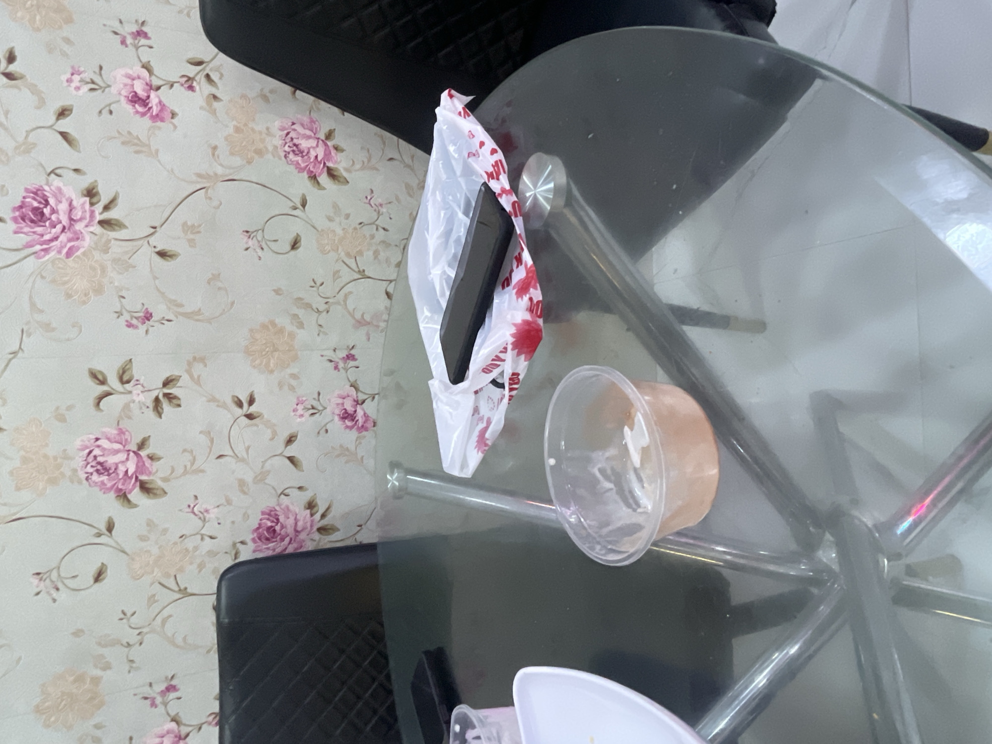 A black smartphone lies on top of a white plastic bag with red lettering on a glass-topped table. To the left of the phone and bag, a clear plastic cup with some remaining pink liquid sits on the table. A white plate is partially visible on the far left side of the table. Two black chairs are visible behind the table. The wall behind the chairs is decorated with a floral wallpaper featuring pink and beige flowers. The scene is indoors, likely in a dining area. The provided information does not specify the exact location within Jalingo, Nigeria.
