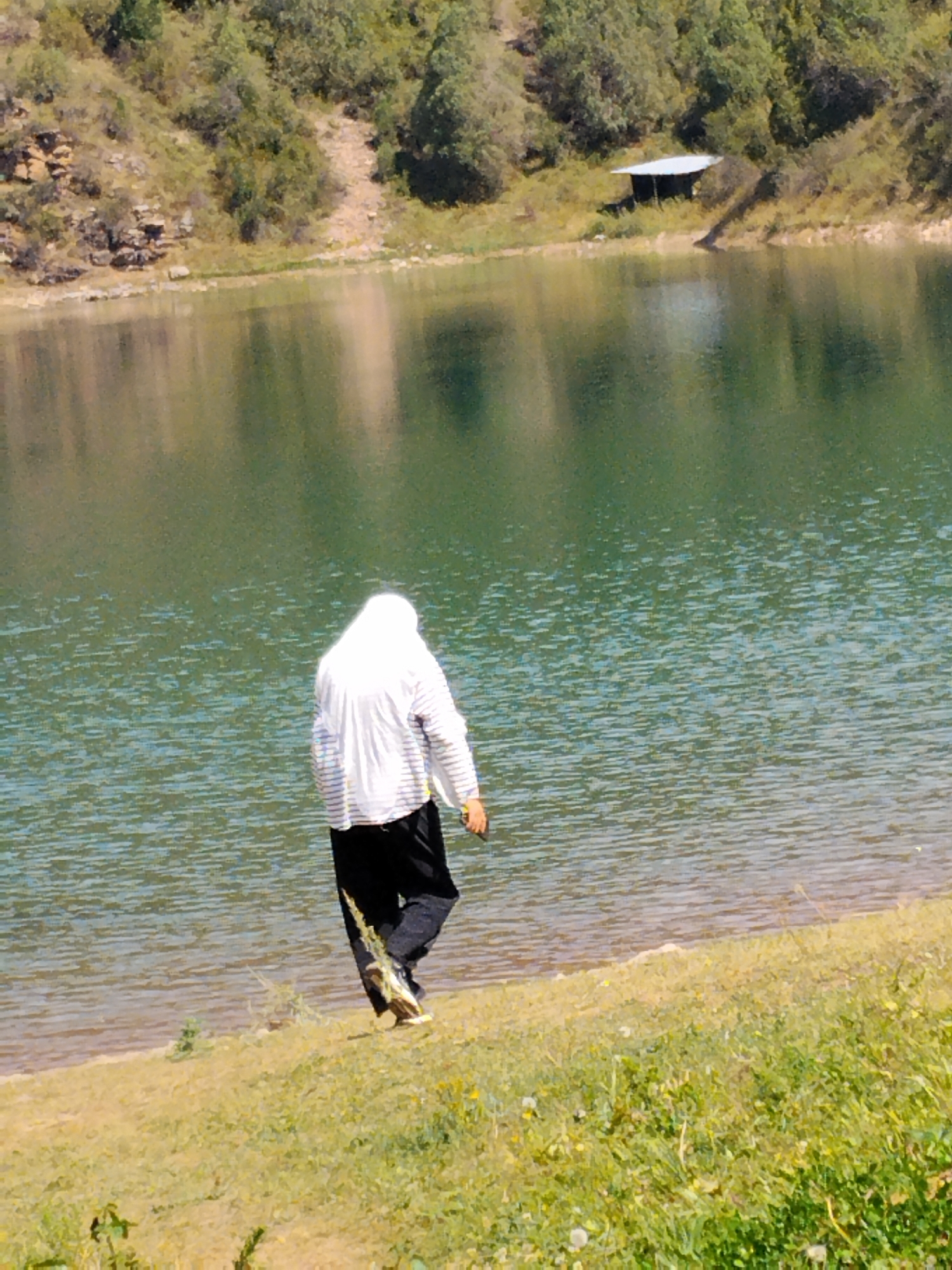 A person wearing a white headscarf and a striped shirt walks on the grassy shore of a lake in Ala-Buka, Kyrgyzstan. The lake's water is a greenish-blue color, reflecting the surrounding trees and hills. In the background, a small, simple structure with a white roof is visible on the hillside. The terrain is generally hilly and covered with vegetation.