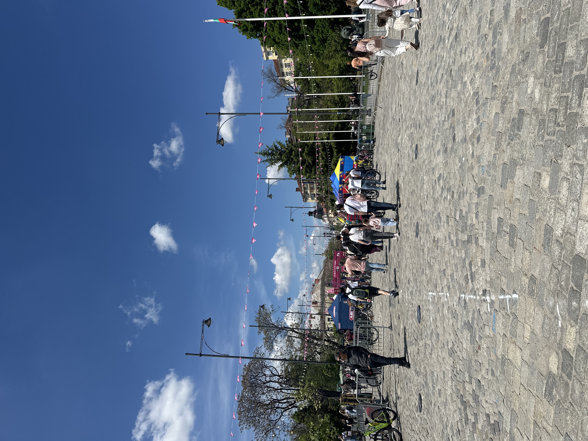 The image captures a street scene in Sofia, Bulgaria, likely during a public event. Cobblestones pave the street where several people stand and walk amidst rows of bicycles, suggesting a cycling-related event or gathering. Metal barriers partition the area, possibly to control pedestrian or bicycle traffic. Above, a series of streetlights are decorated with pink pennant flags, lending a festive atmosphere to the scene. The sky is blue with scattered clouds, and a Bulgarian flag flutters from a pole in the distance. Buildings and greenery line the street, indicating an urban setting within Sofia.