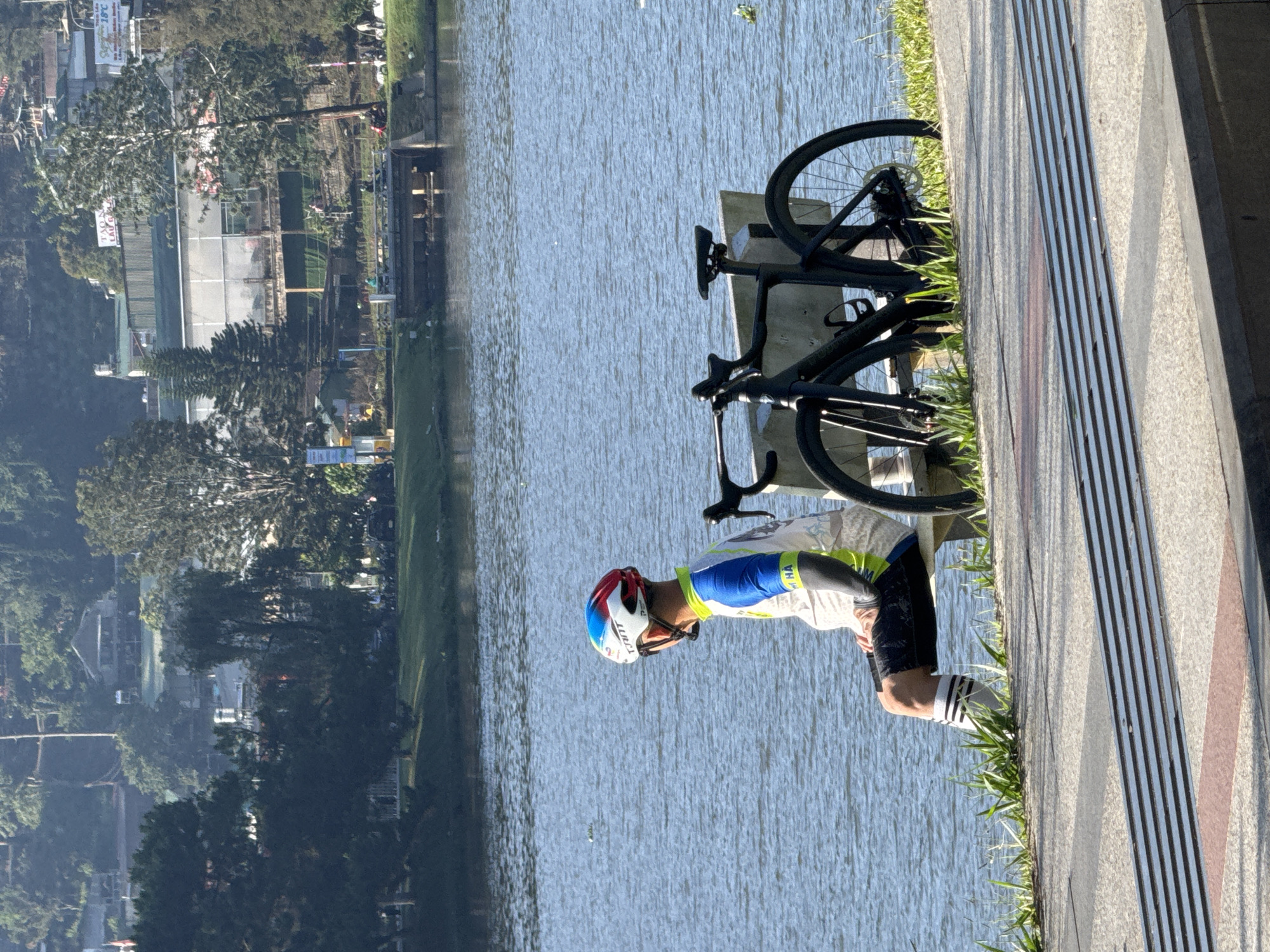 A medium shot captures a cyclist stretching near a black bicycle parked on a sidewalk in Da Lat, Vietnam, with Xuan Huong Lake as the backdrop. The cyclist is wearing a colorful helmet, a light-colored jersey with blue and green accents, dark shorts, and striped socks. The bicycle is leaning against a bench adjacent to the sidewalk, which is decorated with contrasting gray and dark red stripes. Beyond the lake, the cityscape of Da Lat is visible, with buildings and lush greenery. The sky above appears clear.