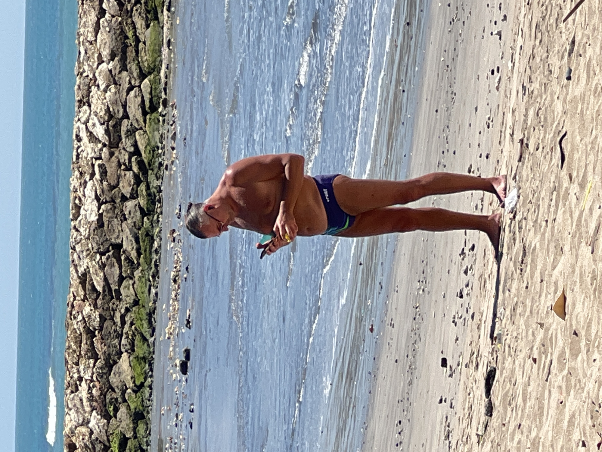A man stands on a sandy beach, looking down at his smartphone. He is wearing dark blue swim trunks with a green stripe. The ocean is behind him, with waves lapping at the shore. A rock jetty is visible in the background. The sky is clear and blue. The scene appears to be a sunny day at the beach.