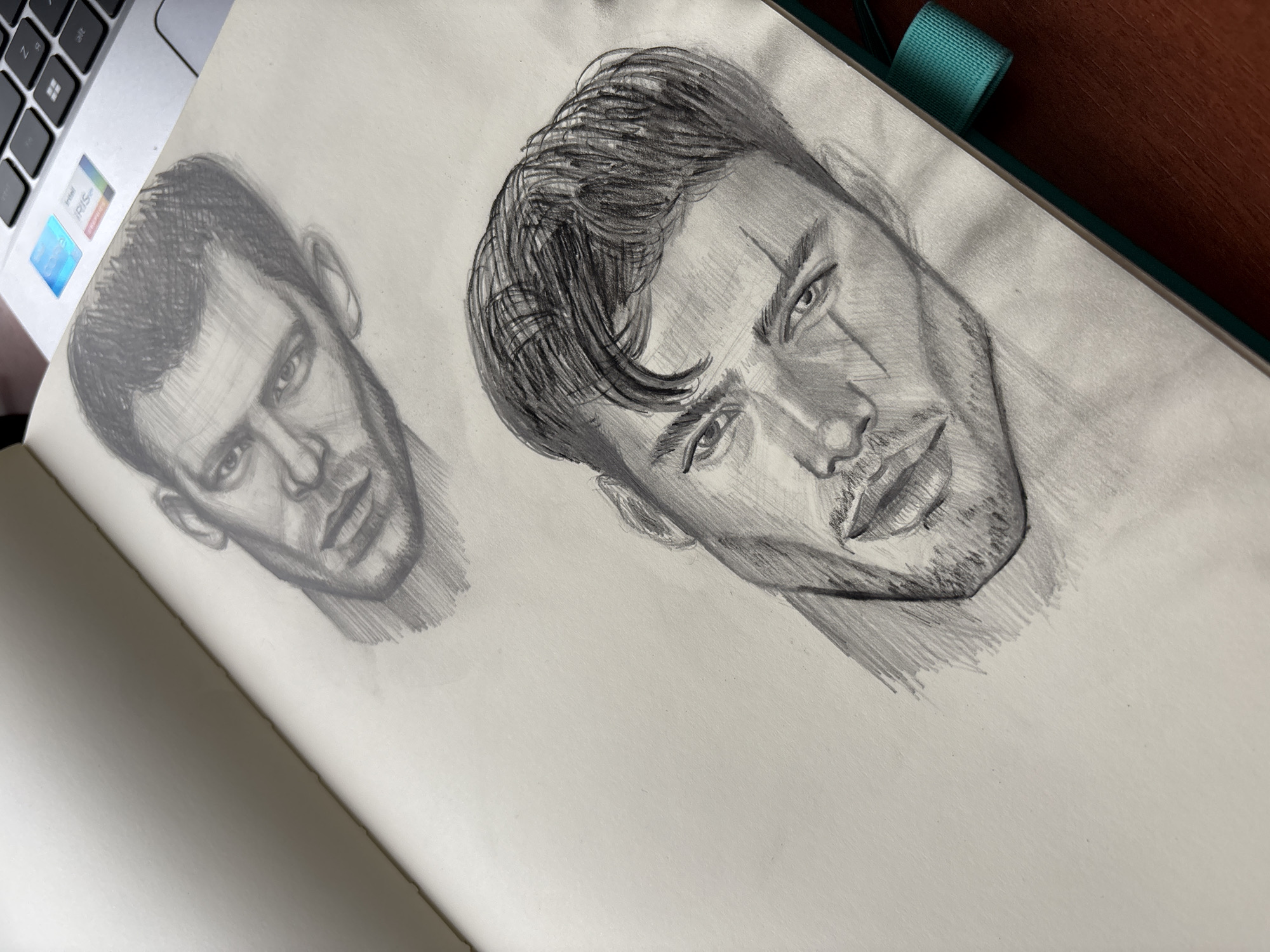 The image shows a sketchbook with two pencil sketches of the same man's face. The sketch on the right is more detailed, with expressive shading, defined hair with a prominent wave, and a scar visible below the left eye. The sketch on the left is less defined. The background includes a laptop with visible keyboard keys and a teal ribbon bookmark. The sketchbook is open and lying on a dark wooden surface. The location in Izhevsk, Russia is not identifiable from the image.