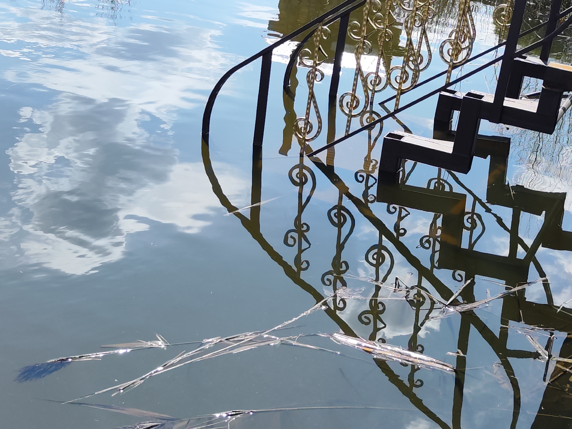 The image depicts a set of stairs leading into water, likely a river or lake, in the city of Chaikovsky, Russia. The stairs have black metal railings with decorative golden swirls. The steps themselves are also black. The water reflects the sky, including blue sky and white clouds. There are some pieces of broken ice or debris floating in the water near the stairs. The photograph is taken at a low angle, emphasizing the water and the reflection of the stairs.