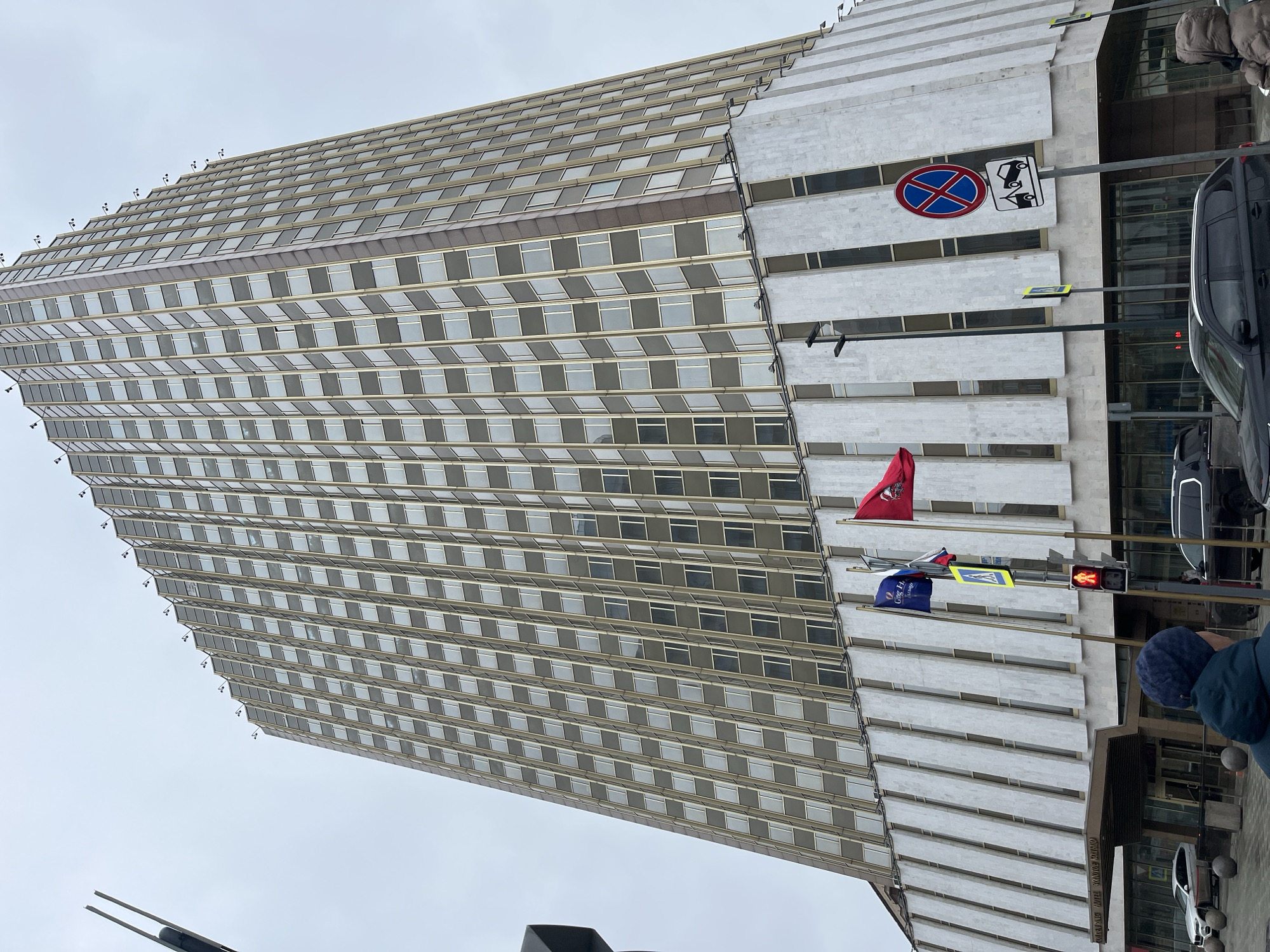 The image depicts the Radisson Collection Hotel in Moscow, Russia. The photograph is taken from a low-angle perspective, emphasizing the height of the building. The hotel is a high-rise structure with numerous windows and a distinctive facade featuring vertical, protruding concrete elements.
There are street signs present, including a "no stopping" sign. Flags are visible, one red and one blue. A traffic light is showing a red light. Pedestrians are visible in the foreground, and cars are parked alongside the building. The sky is overcast.