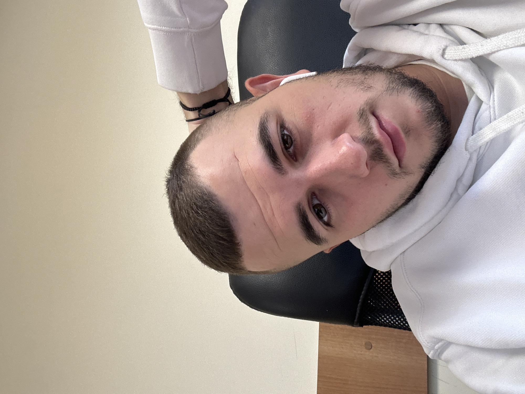 A man is indoors, sitting in a black chair. He is wearing a white hoodie. He has short, dark hair and a beard. He has a bracelet on his left wrist and an earpiece in his left ear. The background consists of a light-colored wall and a wooden surface. The location is not discernable.
