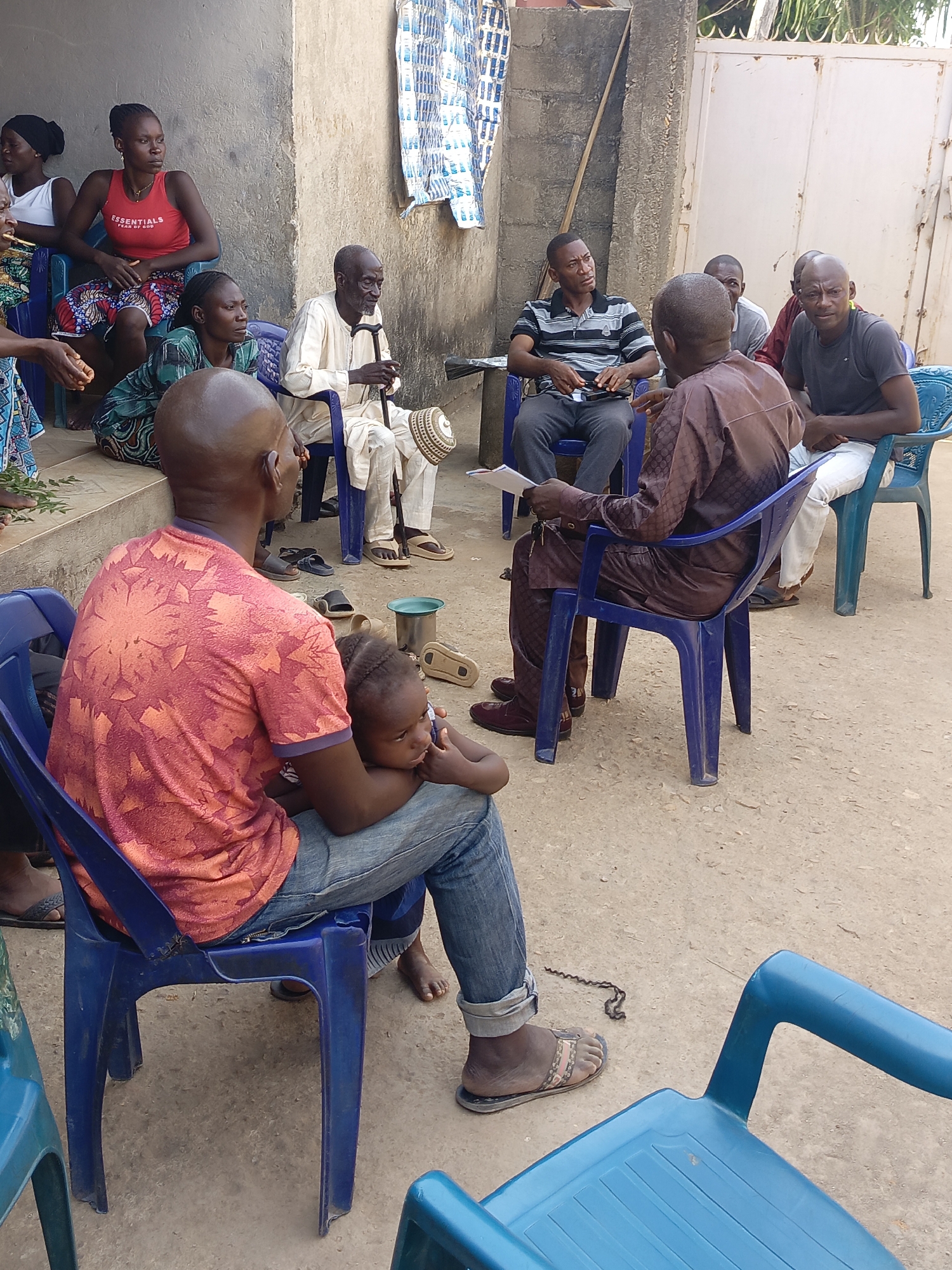 The image shows a group of people gathered in an outdoor space in Akwanga, Nigeria. Nine adults and one child are visible, seated on plastic chairs arranged in a semi-circle. A man in a red patterned shirt sits with a small child on his lap. Another man, dressed in brown traditional attire, is holding papers and appears to be addressing the group. Several other men are listening attentively. A woman in a red tank top is seated on a higher step, along with other women. A senior man in white traditional clothing sits with a walking stick. A curtain hangs from the building. The setting includes a concrete floor and a metal gate. The ground is dry, with a few scattered objects.