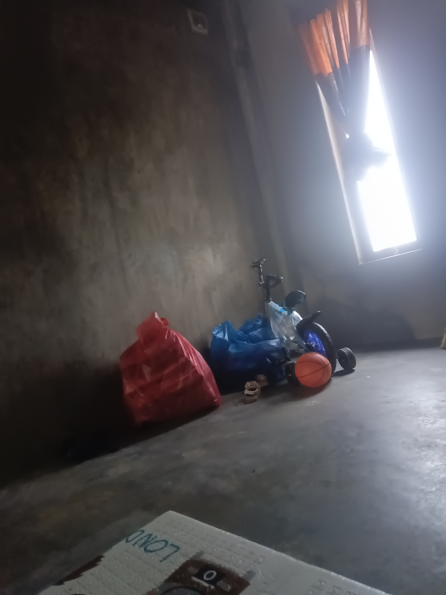 An indoor scene depicts a concrete floor and a dark gray wall. On the floor, a red plastic bag and a blue plastic bag are visible, along with a toy tricycle and an orange basketball. A window with orange curtains is visible on the right side of the image, letting in bright light. A white object with some text is also visible in the foreground. The scene is set in a location identified as Unknown, Unknown.