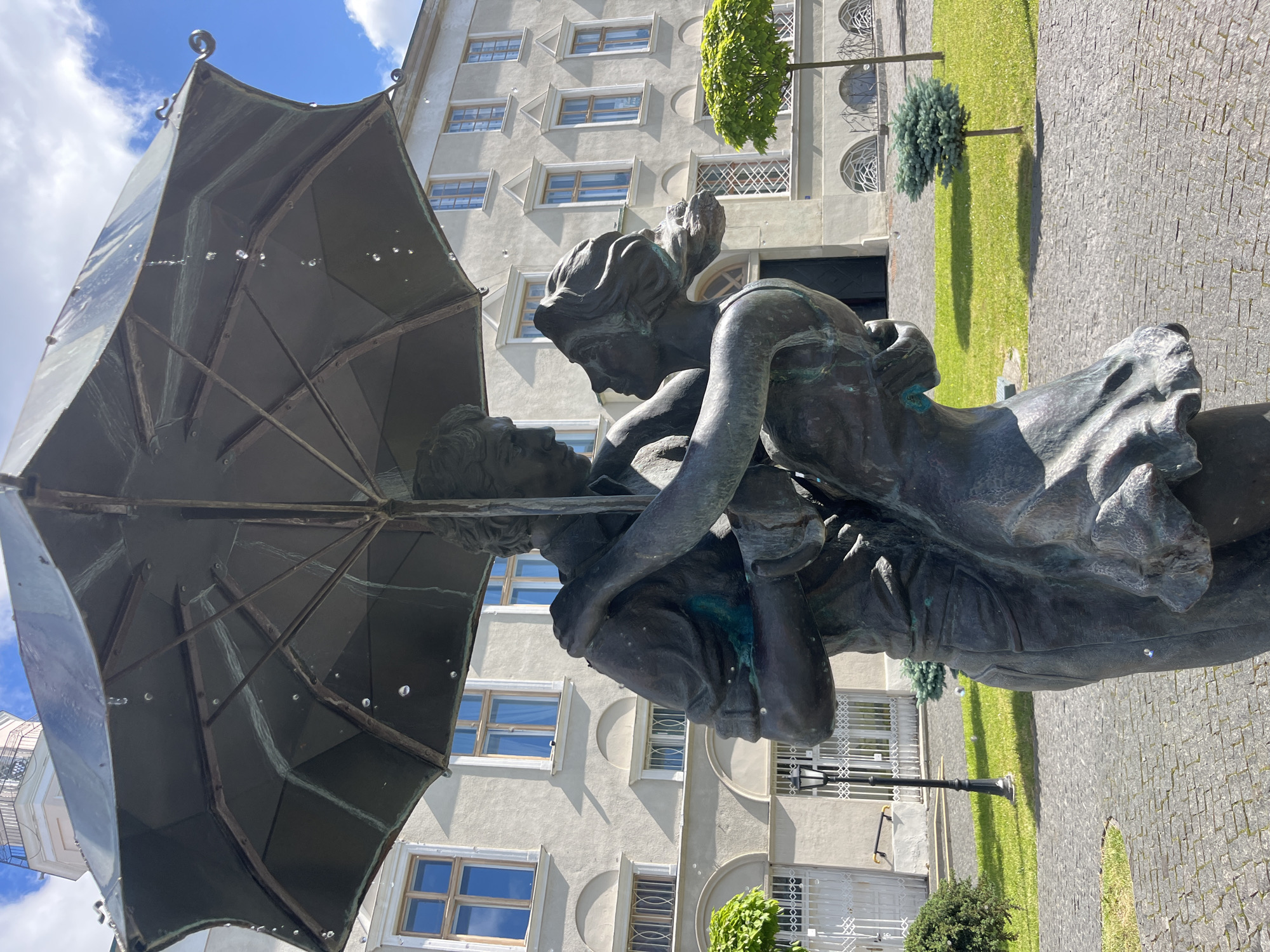 This image shows a bronze sculpture of a couple embracing under an umbrella, located in the city of Drohobych, Ukraine. The sculpture appears to be a fountain, as water droplets are visible falling from the umbrella. The couple is depicted in a close embrace, with the man holding the woman and the umbrella. The background features a light-colored building with arched windows and a tower-like structure. Green grass and cobblestone pavement surround the sculpture.