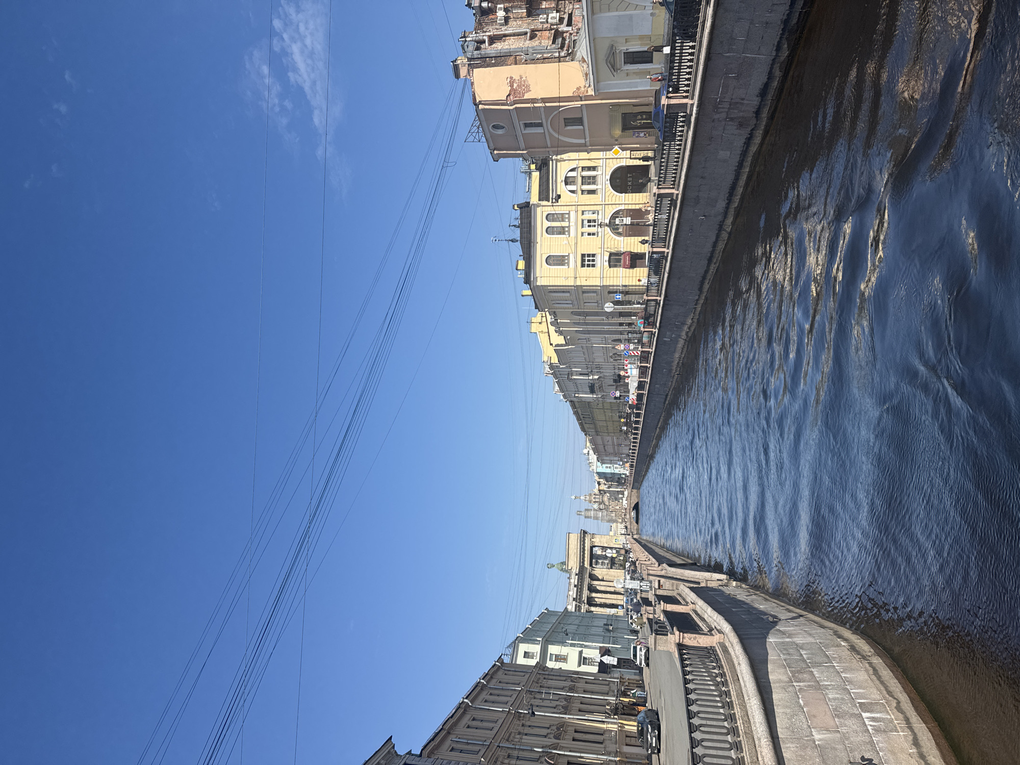 The image displays a sunny cityscape featuring a wide canal, flanked by buildings and embankments, likely in Saint Petersburg, Russia. The perspective looks down the length of the canal.
On the right side of the image (when oriented correctly), the canal's dark blue water is rippled, reflecting sunlight in shimmering streaks. A concrete or stone embankment with a dark metal railing runs alongside the water. Beyond this, a continuous row of multi-story, historic-style buildings lines the canal. These buildings exhibit various architectural details, with facades in shades of yellow, light brown, and grey. Some buildings show signs of age, with exposed brickwork or air conditioning units mounted externally. One prominent yellow building features arched windows and a circular sign or emblem, below which faint text "ROCKET" is visible. Traffic signs and streetlights are scattered along the street in front of these buildings.
On the left side of the image, another embankment, also with a metal railing, parallels the canal. A paved roadway runs along this bank, where several cars are visible, some parked and others possibly in motion. Further left, another line of substantial, classical-style buildings stretches into the distance. A person on a bicycle can be seen on the left embankment in the foreground.
The sky overhead is a bright, clear blue with a few scattered, wispy white clouds. Numerous overhead wires crisscross the sky, particularly on the left side of the frame, suggesting urban infrastructure.
The scene depicts a calm, bright day. The strong sunlight and clear shadows indicate it is likely late morning or early afternoon. The overall impression is one of a historic European city's urban waterway on a pleasant day.