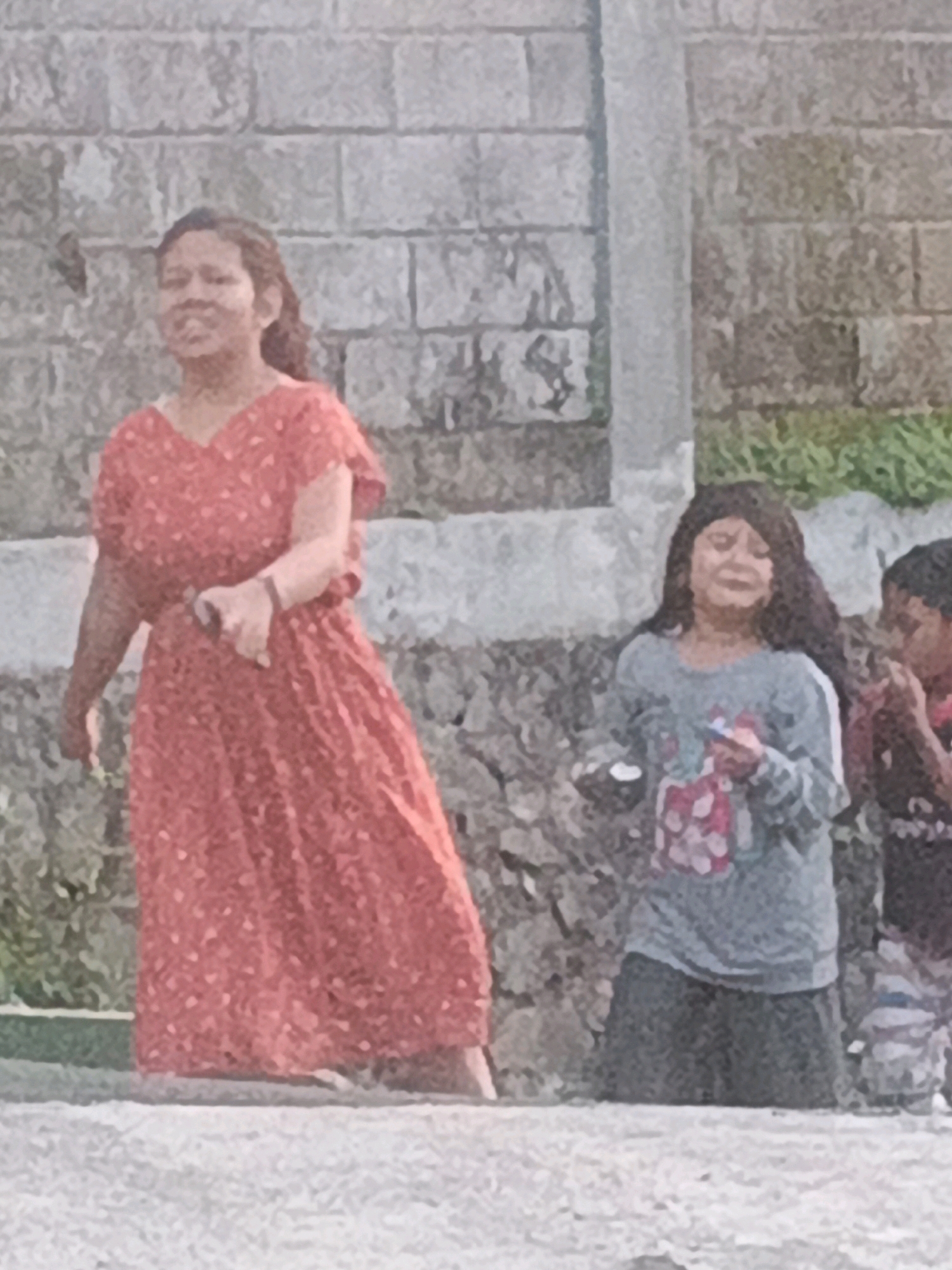 In Keji, Indonesia, a group of individuals stand outdoors against a textured brick wall. The woman on the left is wearing a red, patterned dress and appears to be speaking or singing. To her right, a young girl in a grey long-sleeved shirt and dark pants is holding a small object, possibly a toy or candy. Another child, partially visible on the far right, is wearing a striped shirt and shorts. The ground in front of them is a light-colored, textured surface, and behind them, lush green foliage is visible. The scene suggests a casual outdoor gathering or playtime.