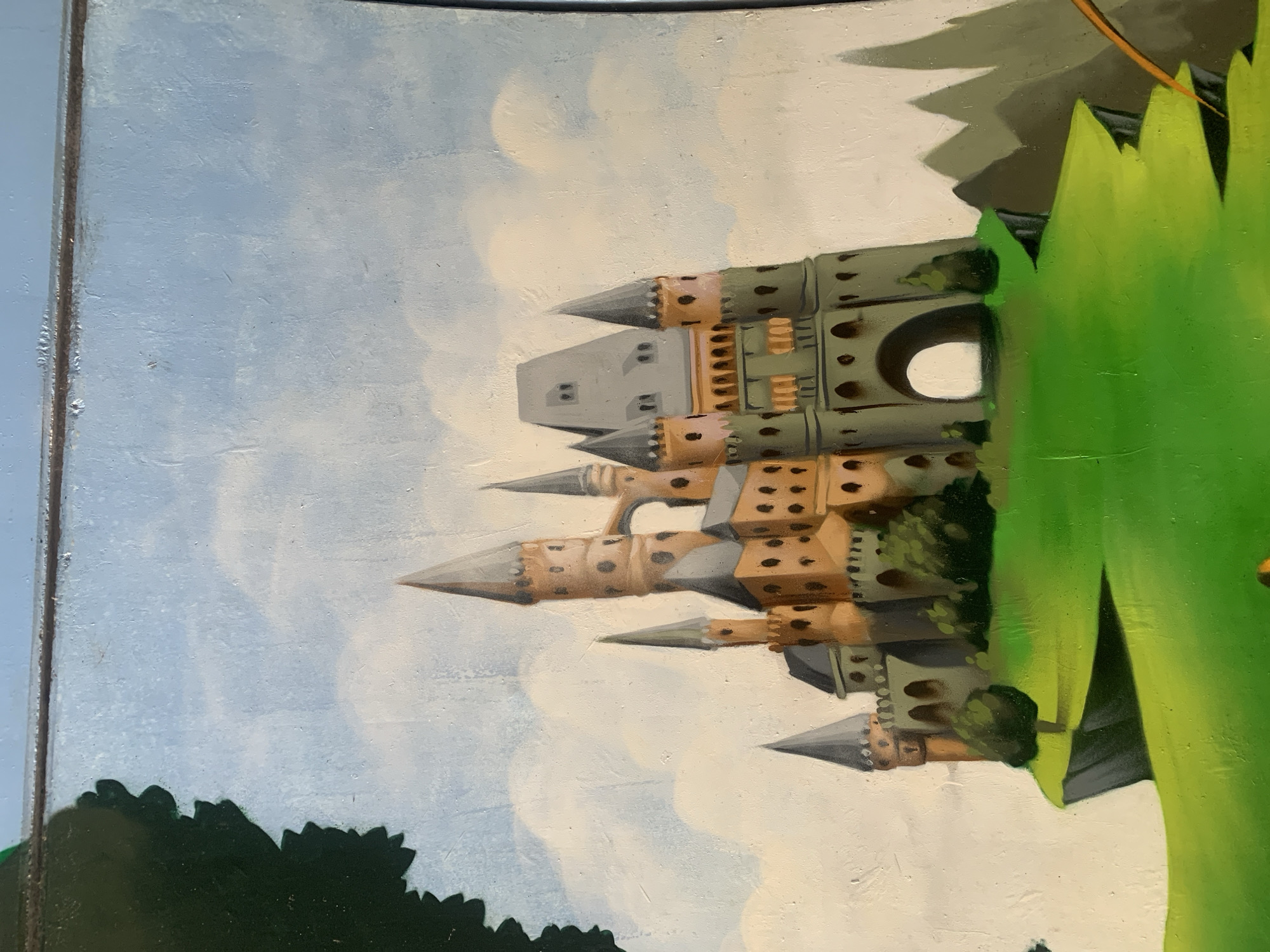 The image depicts a painting of a fantastical castle perched on a cliff overlooking a vibrant green landscape. The castle is elaborate, featuring multiple towers with conical roofs, arched windows, and various architectural details in shades of brown, gray, and green. The sky above is a light blue with soft white clouds. In the foreground, the green hill slopes downward, with stylized trees and rocky outcrops. A large, dark green, leafy silhouette is visible on the far left edge of the image, suggesting a partial view of a tree or foliage. The overall style of the painting is illustrative and whimsical. There is no discernible location within the city of Odivelas, Portugal depicted in this artwork.