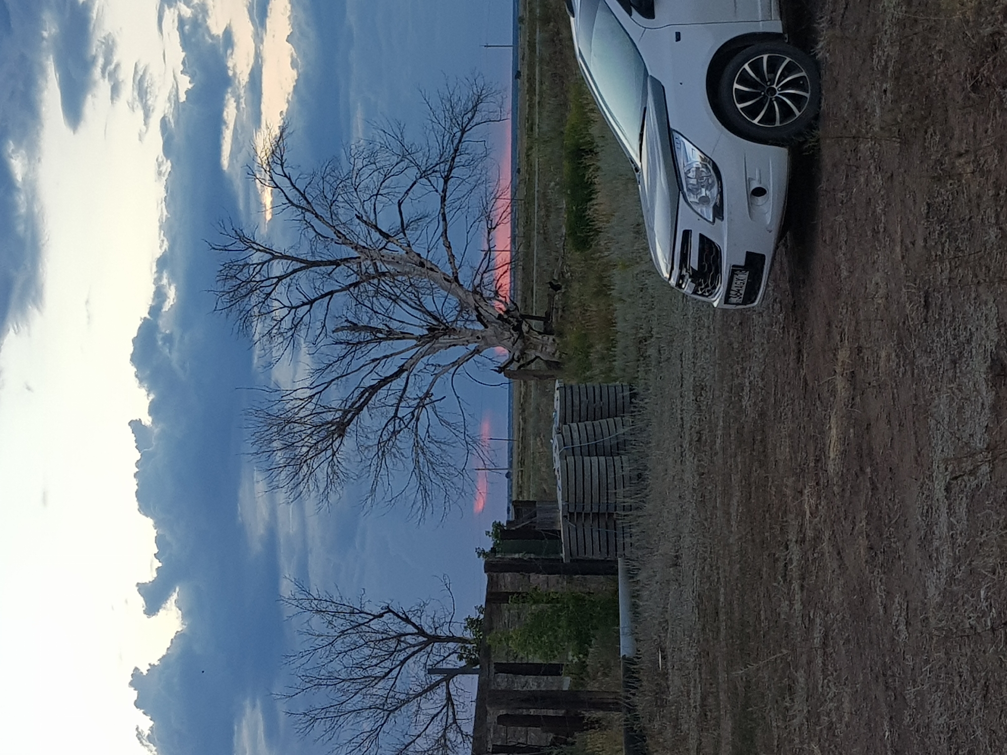 The image shows a white Chevrolet Cobalt parked on a dirt road in a rural area. The car is facing right and is partially visible in the foreground. Behind the car, there is a landscape with dry grass, a few bare trees, and some wooden structures. In the distance, there is a flat horizon with a cloudy sky. The sky has a mix of dark blue and light gray clouds, with a hint of pinkish-orange light visible on the horizon, suggesting either sunrise or sunset. There are no people visible in the image. The location is identified as the village district of Dolen, Kazakhstan.