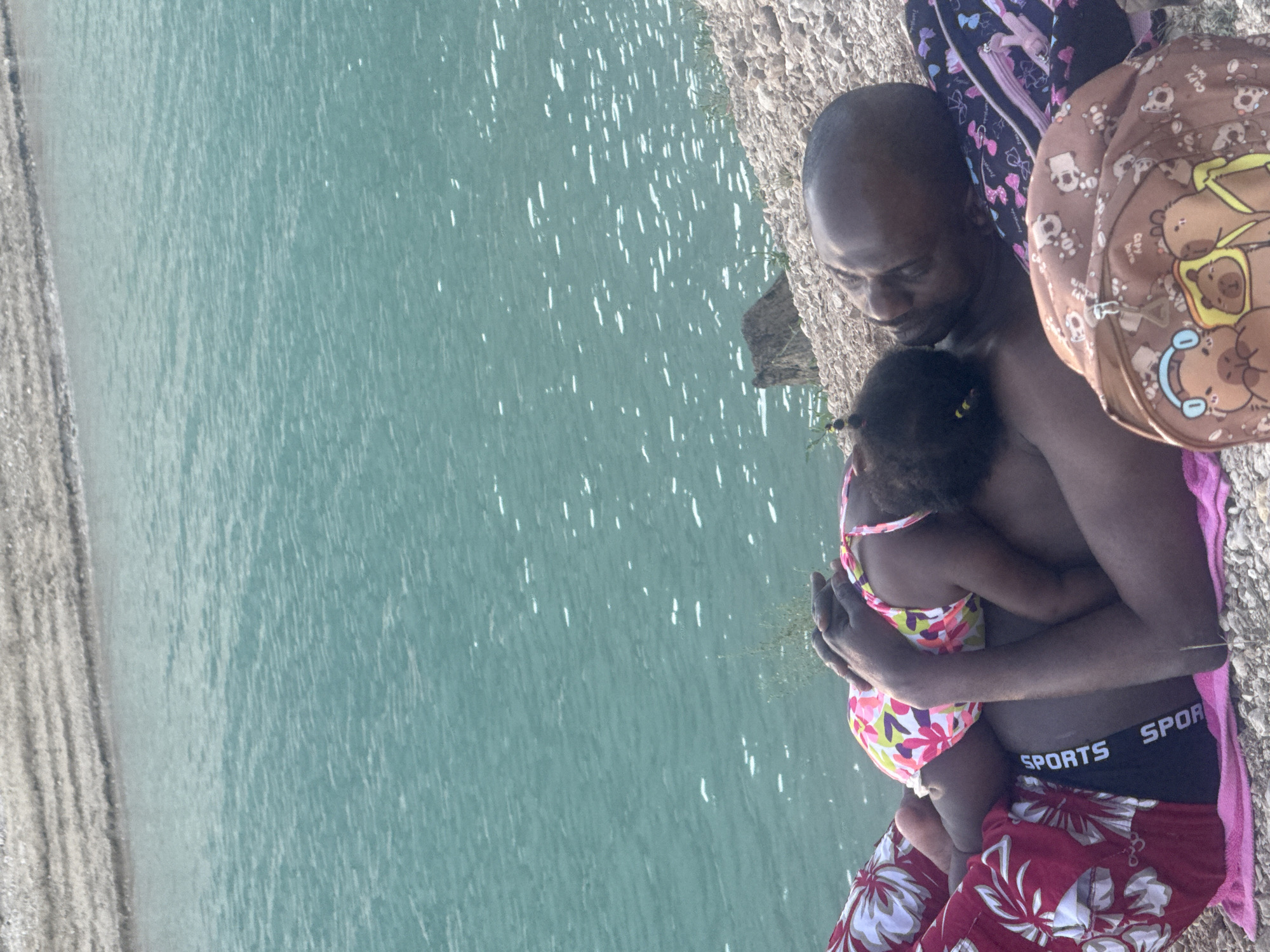 In Maisod, France, a man and a young child lie on a rocky shore next to a body of turquoise water. The man is lying on his back, and the child is asleep on his chest, wearing a colorful swimsuit. The man has his arms around the child, and both appear relaxed. The background shows more of the rocky shore and the expansive water. Two backpacks are also visible near the man. The scene suggests a peaceful moment of rest or bonding by the water.