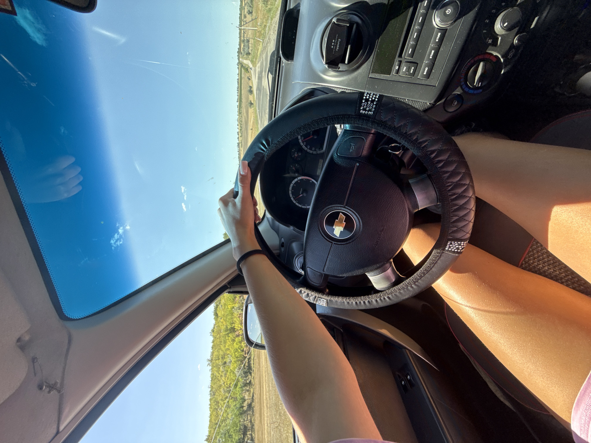 The image is taken from inside a car, looking forward through the windshield. A person's hands are visible on the steering wheel. The exterior view shows a rural landscape with a road stretching into the distance under a clear blue sky with scattered clouds. The car's dashboard, including the steering wheel, instrument cluster, and radio, is visible. The lighting suggests it is daytime, likely sunny. No specific details identify the location as Basarabeasca, Moldova.