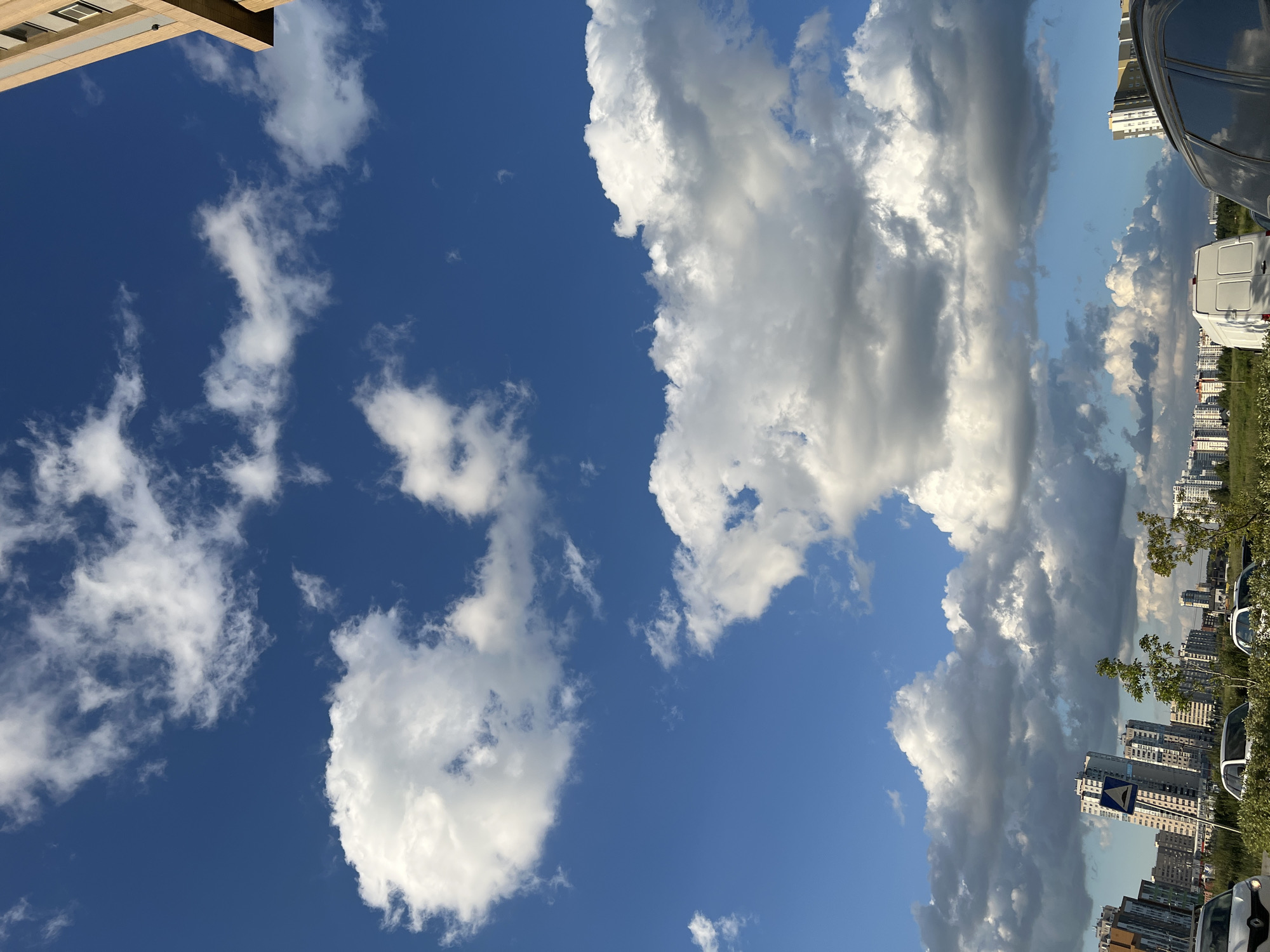 This media file depicts a view of the sky with scattered clouds, with a cityscape and parked vehicles visible in the background. The scene appears to be outdoors on a sunny day, with a bright blue sky dominating the upper portion of the image. The clouds are predominantly white and fluffy, suggesting fair weather, though some darker, more ominous clouds are present towards the right side of the frame, hinting at potential changes in weather.
The cityscape in the background features a collection of multi-story buildings, likely apartment complexes, with varying architectural styles and colors. The buildings are densely packed, suggesting an urban or suburban environment. Several vehicles are parked along what appears to be a street or parking area, including a white van and parts of other cars.
The time of day appears to be daytime, given the bright sunlight and blue sky. The presence of a building with windows and balconies in the upper left corner suggests an inhabited area. The overall mood conveyed by the image is one of a typical day in a city, with a focus on the sky and its atmospheric conditions.
There is a visible traffic sign in the background, a blue diamond shape with a white arrow pointing downwards and to the left, indicating a direction. No people are visible in the frame. The location context provided is Yekaterinburg, Russia, which aligns with the appearance of the buildings and urban landscape.