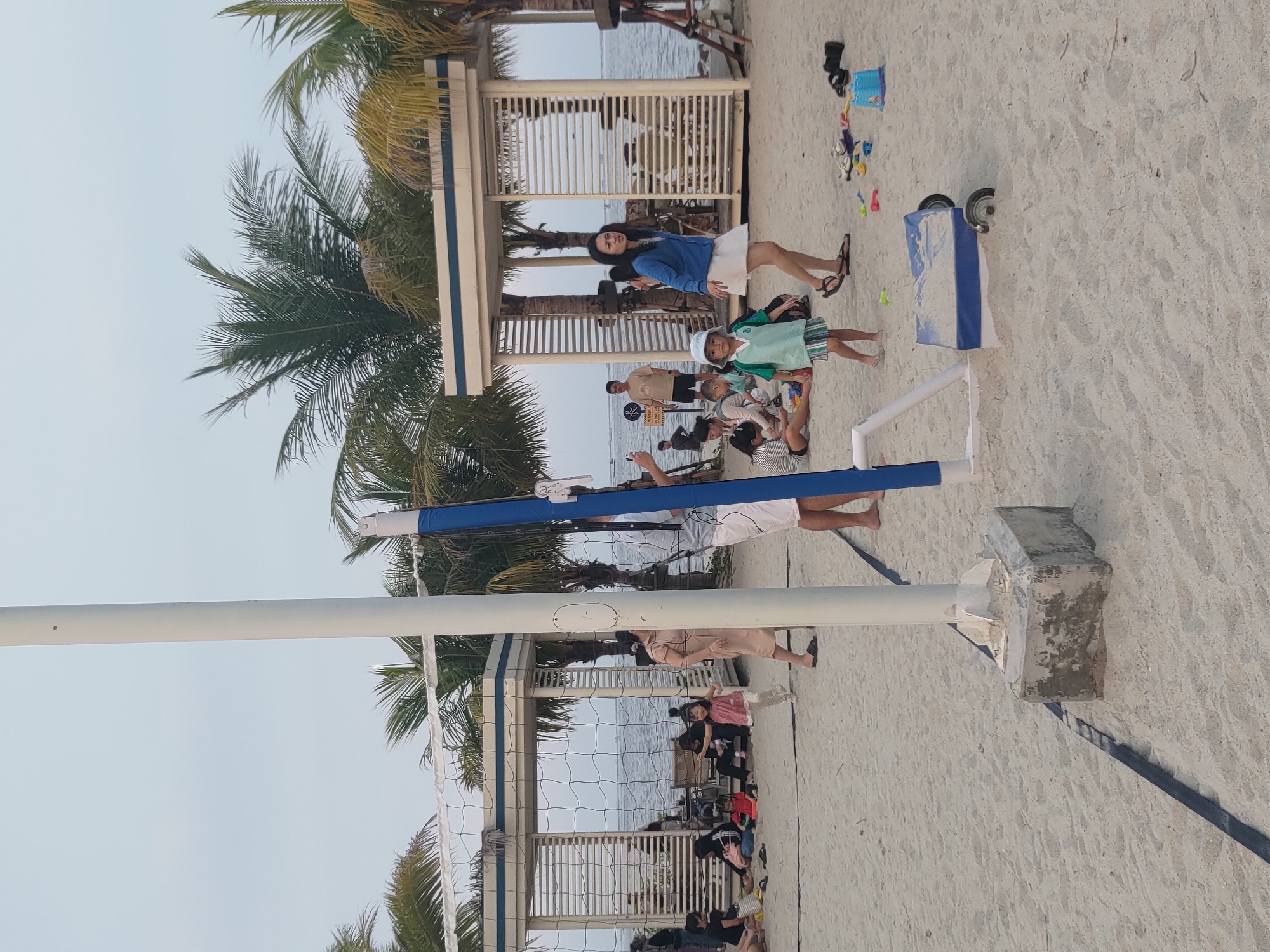 The image depicts a beach scene in Kosambi Barat, Indonesia, with a volleyball net and several people scattered across the sand. A white pole, part of the volleyball net system, stands prominently in the foreground, with a concrete base. To the right of the pole is a blue and white cart with wheels, filled with sand. Palm trees line the background, and a body of water can be seen beyond the net. Several individuals, including adults and children, are engaged in various activities such as sitting, playing, and walking on the beach. A woman in a blue cardigan and white skirt walks past the cart, while a child in a green and white striped shirt stands nearby. Other people are seated in the shaded areas under the structures behind the net. The sand appears light in color and scattered with debris. The sky is overcast.