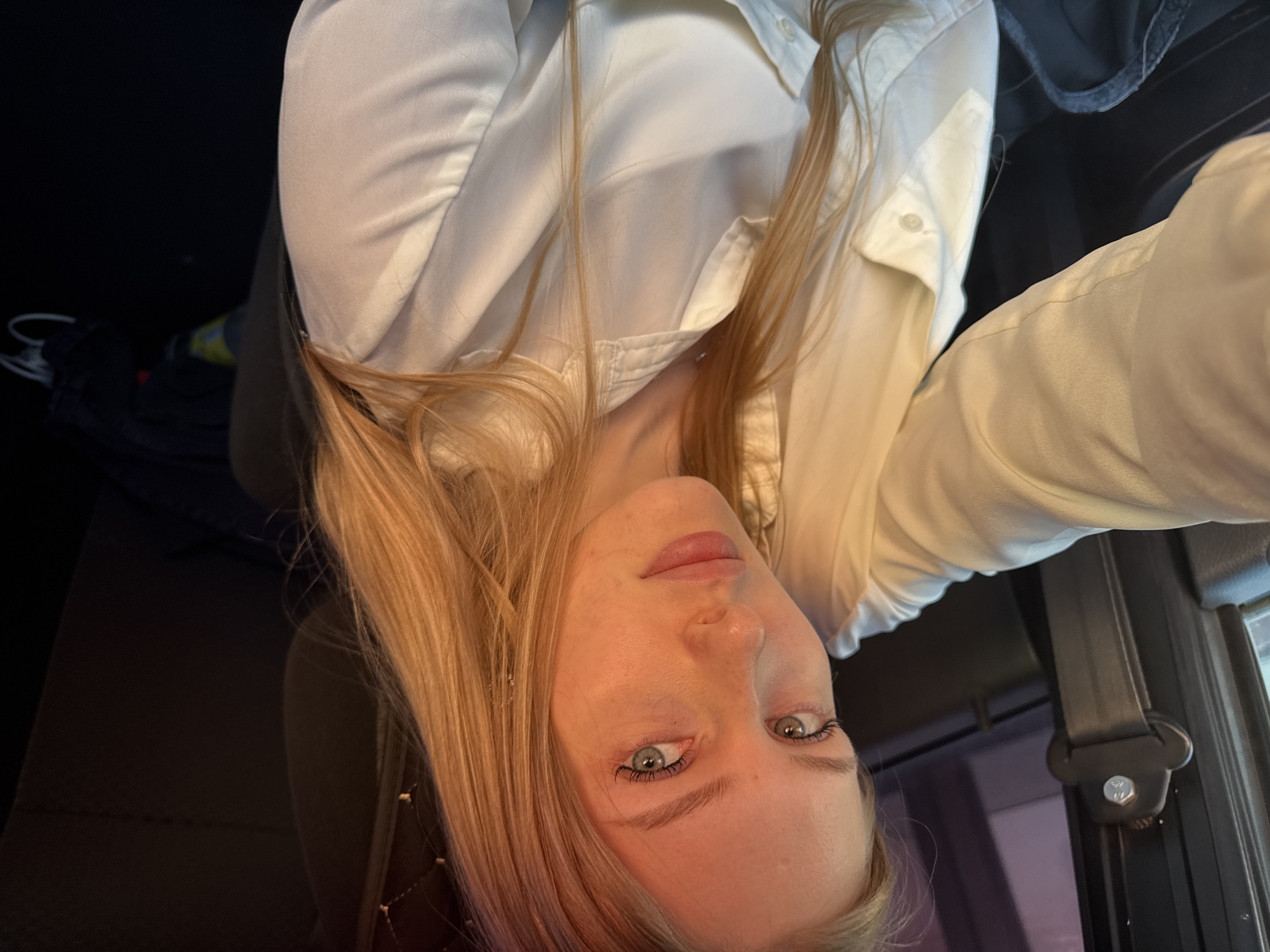 A fair-skinned woman with light blonde hair and green eyes is taking a selfie. She is wearing a white collared shirt. She is seated in a car with a seatbelt visible. The background is dark, and the specific location within the city of Perm, Russia cannot be determined from the visual information provided.