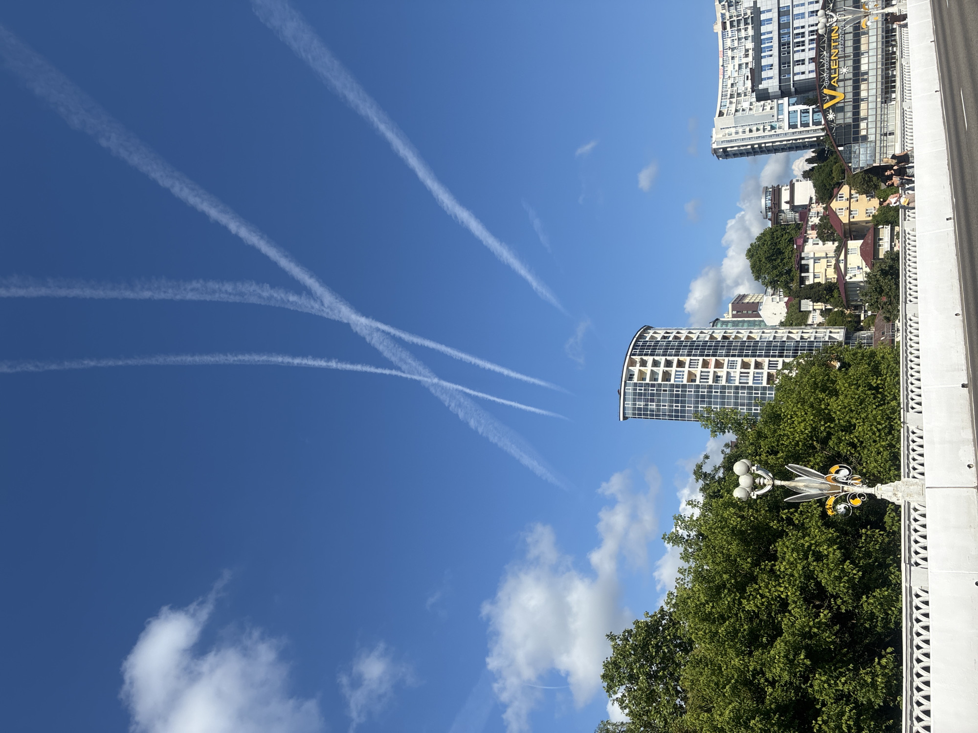 The image captures a view from a bridge in Sochi, Russia, looking towards a cityscape with several modern high-rise buildings. The sky is a vibrant blue, with scattered white clouds and multiple distinct contrails from aircraft, forming a pattern of intersecting lines. In the foreground, a white bridge railing with intricate decorative patterns is visible, alongside a lamppost with a unique design. Dense green trees line the left side of the frame, partially obscuring some of the buildings. Several people are seen walking on the bridge in the mid-ground, some appearing to be tourists. The buildings in the background vary in architectural style, with one prominent curved skyscraper being a notable feature. A large building with a yellow "VALENTIN" sign is also visible, suggesting a commercial area. The overall scene depicts a sunny day in a coastal city, characterized by both natural elements and modern urban development.