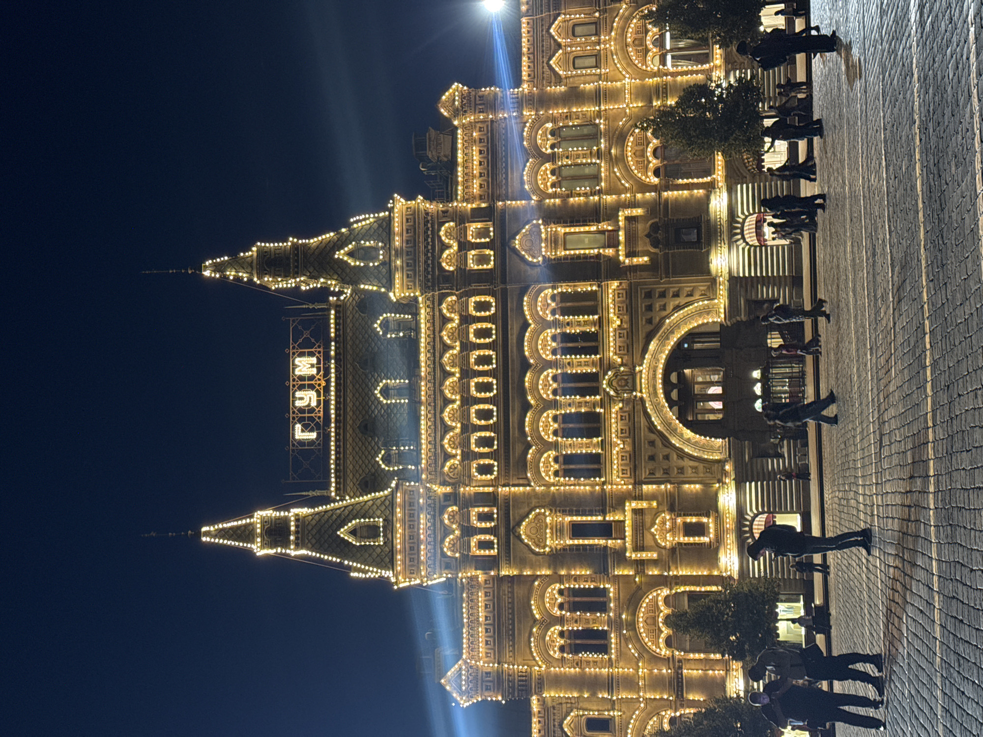 This is a nighttime, eye-level, full-shot image of the GUM department store, located on Red Square in Moscow, Russia. The facade of the building is elaborately decorated with numerous golden lights, which outline its architectural features, including its towers, arches, and windows. The lights create a warm, festive glow against the dark night sky. The cobblestone ground in front of the building is visible, and several people are scattered across it, appearing to be walking or standing, some engaged with their phones. Trees are visible on the sides of the building. The overall impression is of a grand, well-lit historical landmark in a bustling urban environment at night.