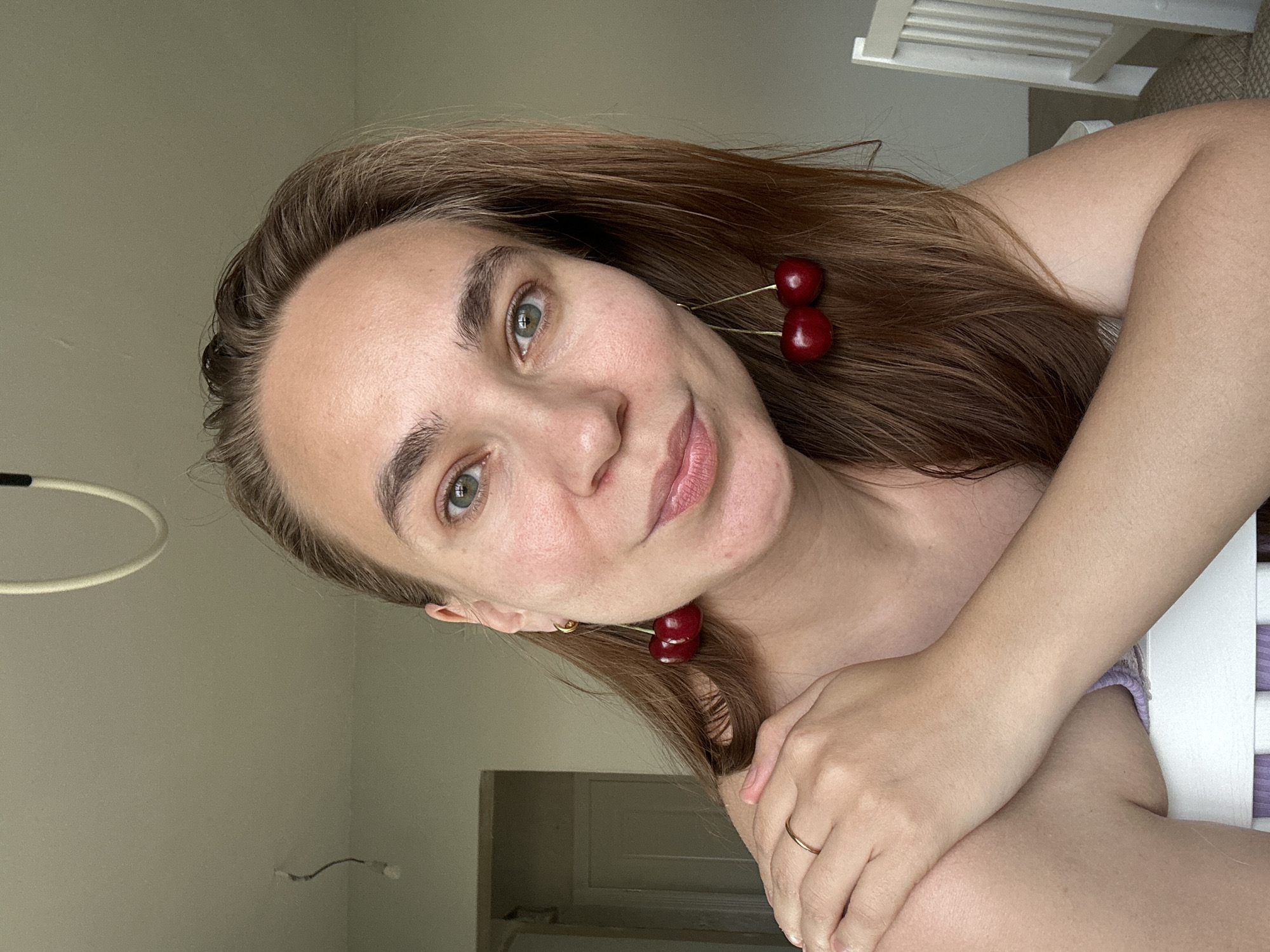 A portrait-style image features a young woman posing indoors in what appears to be a residential room in Kharnavy, Russia. She has long, straight, light brown hair and direct green eyes that engage the viewer. A faint, gentle smile graces her lips. She is wearing distinctive cherry-shaped earrings, with one pair clearly visible near her right ear and another hinting near her left. A simple gold ring adorns the ring finger of her left hand, which is visible resting against her arm or chest.
The woman's skin appears clear and well-lit by soft, natural light, suggesting it is daytime. The background consists of plain, light-colored walls, possibly off-white or light grey, indicating a simple and uncluttered setting. Parts of a white, slatted piece of furniture, possibly a chair back or headboard, are visible in the upper right and lower right corners. There's also a faint wire or light fixture visible on the wall in the mid-left background. No discernible text is visible within the image. The overall mood is calm and inviting.