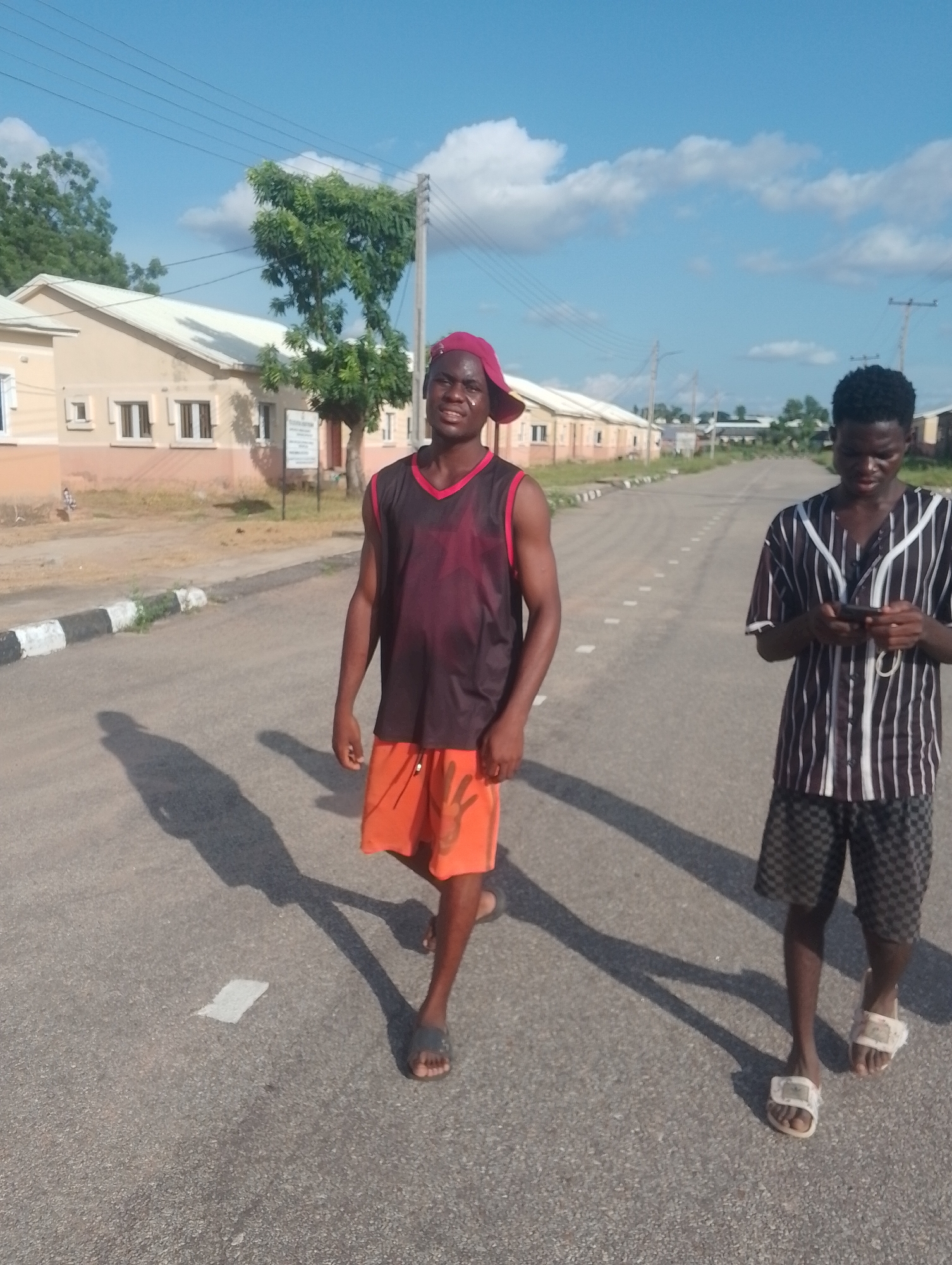 Two young Black males are walking on a paved road in front of a row of beige, single-story buildings. The male on the left is closer to the camera and is wearing a dark sleeveless shirt with red trim, orange shorts, and a red cap. He is looking at the camera and smiling. The male on the right is wearing a striped shirt and checkered shorts. He is looking down at his phone. The road has dashed white lines and the buildings have white roofs and dark windows. There are trees and utility poles in the background. The sky is blue with scattered clouds.