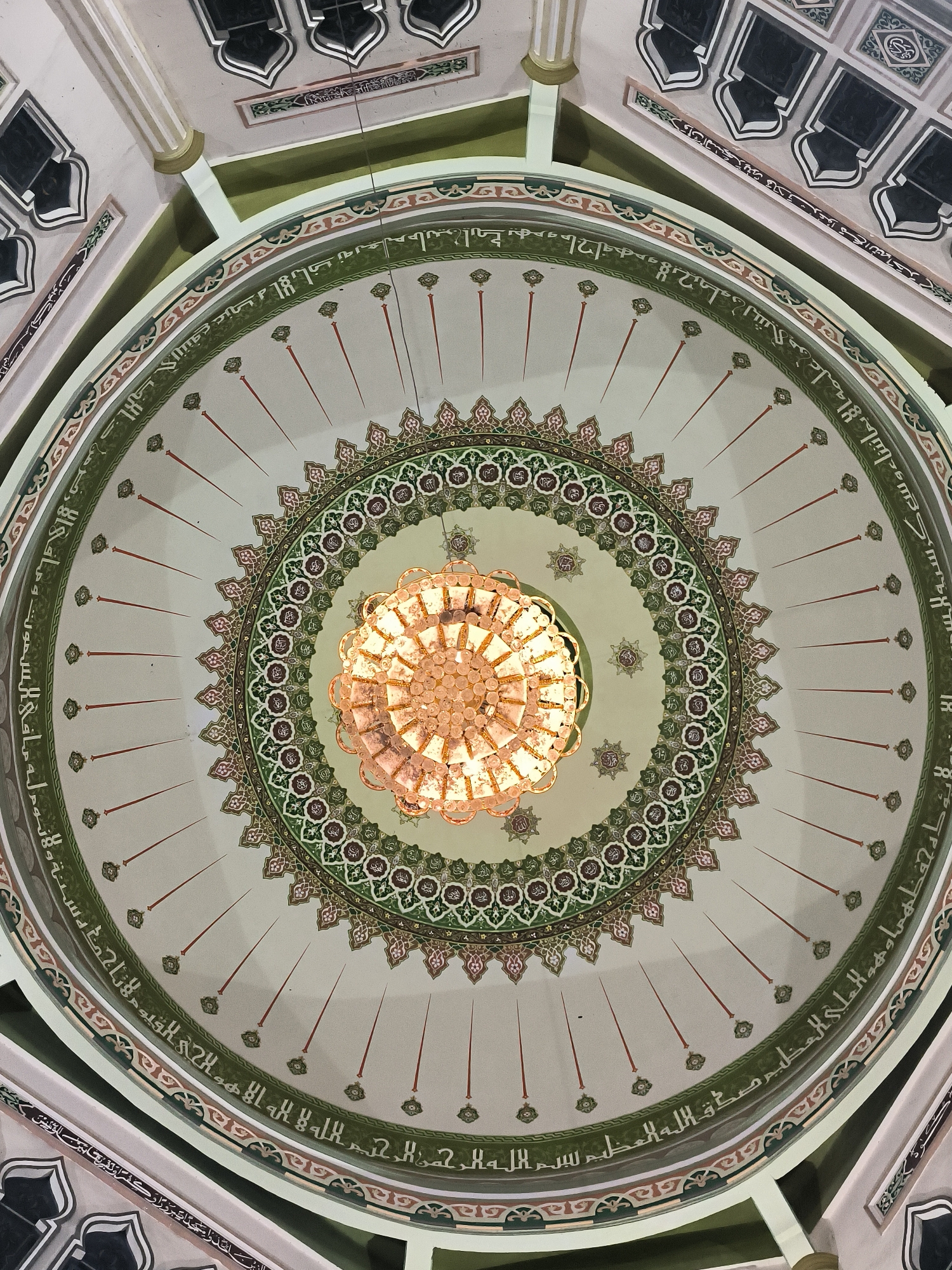 The image shows the interior of a mosque dome in Lhokseumawe, Indonesia. A large chandelier hangs from the center of the dome, which is decorated with concentric circles of intricate patterns and calligraphy. The text is in Arabic and appears to be verses from the Quran. The dome itself is painted in light colors, and the overall impression is one of ornate and religious architecture.