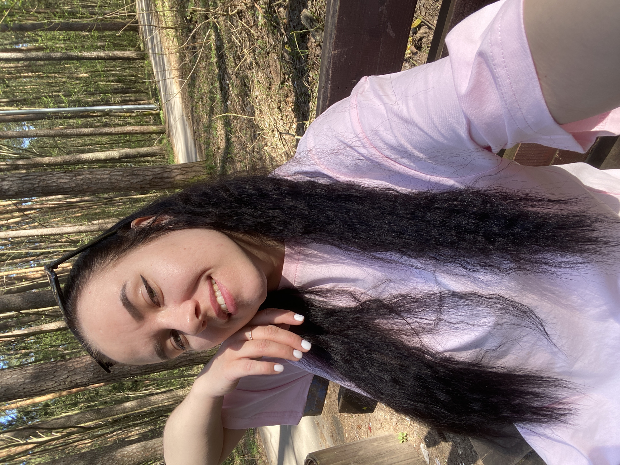 A young woman poses for a selfie in a park in Perm, Russia. She is sitting on a wooden bench, wearing a light pink T-shirt and gold ring. Her long, dark hair flows down her left side. She has fair skin, dark eyebrows, and is smiling. Sunglasses are propped in her hair. The background shows a grove of trees with a path running through it. The ground is covered with fallen leaves. It is a sunny day, and the scene is brightly lit.
