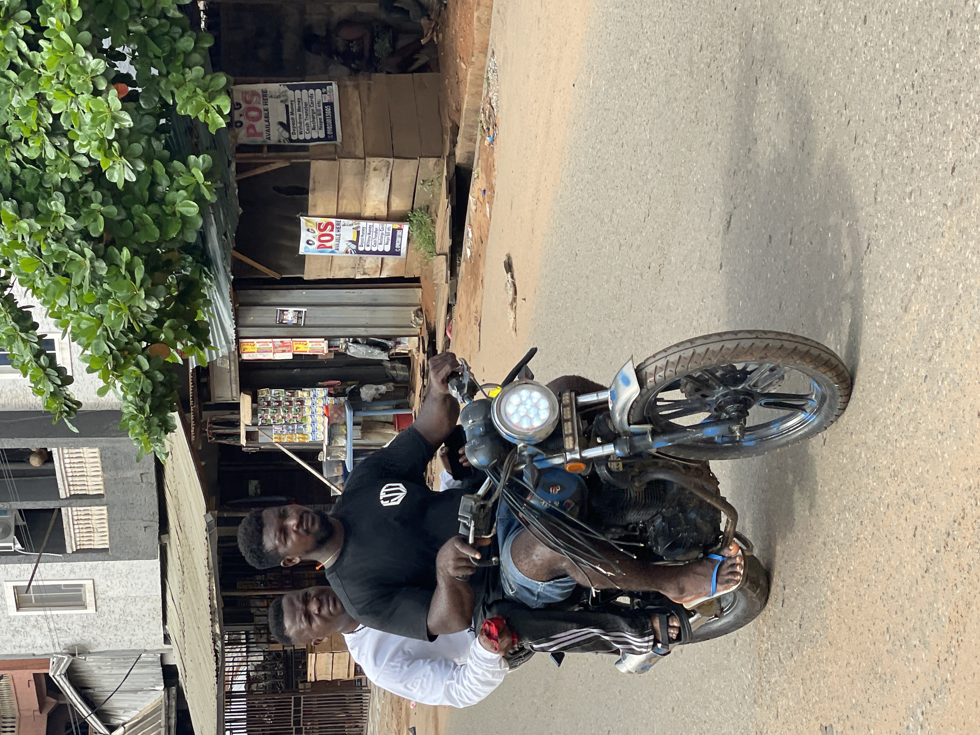Two men are riding a motorcycle on a paved road. The rider is wearing a black t-shirt and denim shorts, with blue flip-flops on his feet. The passenger is wearing a white long-sleeved shirt. The motorcycle has a round LED headlight. In the background, there is a building with advertisements for POS (Point of Sale) services, indicating services such as deposit money, withdraw money, cash transfer, recharge cards, and Nepa Bill payment, with the phone number 09038015805 listed. The location is Nkpor, Nigeria.