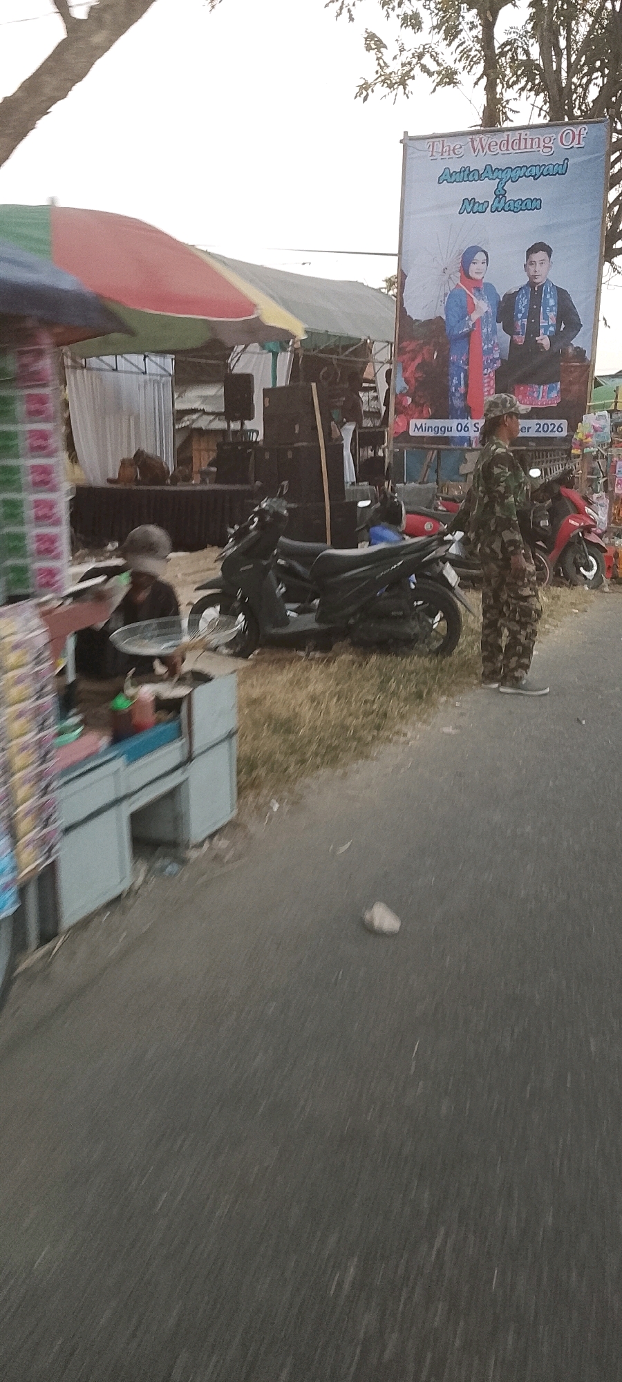 The image depicts an outdoor scene in Sarimukti, Indonesia, likely on a street adjacent to a grassy area. A large banner on the right announces "The Wedding Of Anita Anggriyani & Nur Hasan" and indicates the date "Minggu 06 September 2026". A person in camouflage uniform stands facing away from the camera, looking towards the right of the frame, next to a red motorcycle. In the foreground, to the left, a food stall with colorful packaging is visible, and a person wearing a cap is seated behind it, appearing to be serving or preparing food. Behind the food stall, several motorcycles are parked. Further back, a stage area is set up with speakers and a backdrop, suggesting an event is or will be taking place. The overall lighting suggests it is late afternoon or early evening.