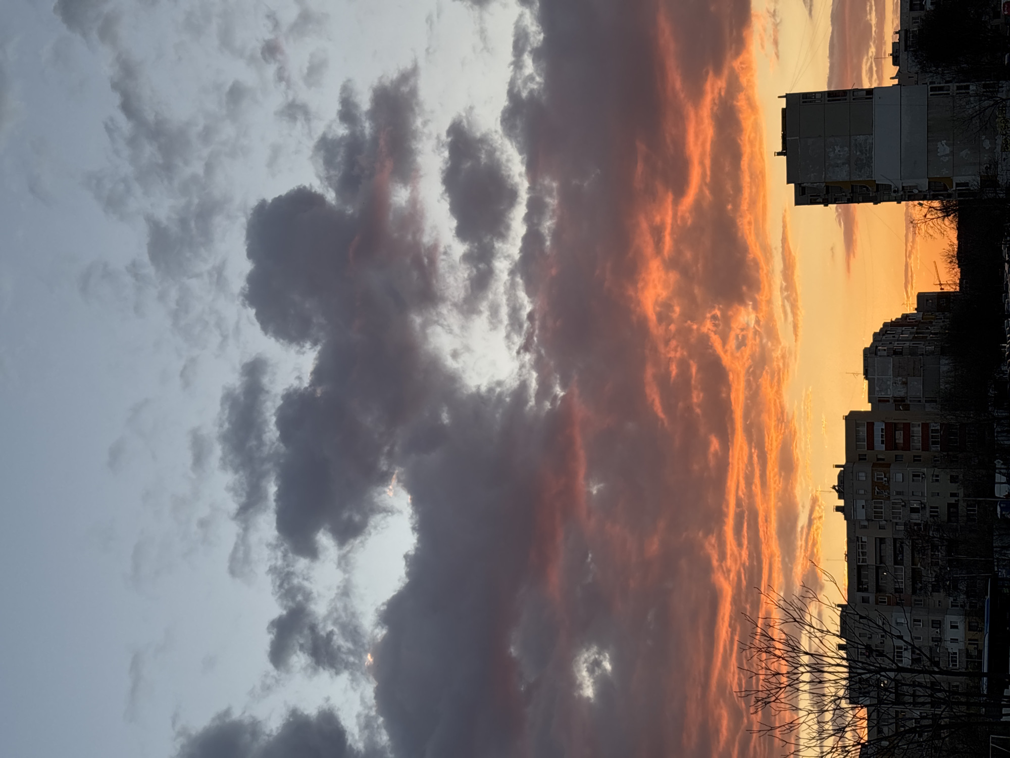 The image depicts a sunset over an urban area. The sky is divided into two distinct parts: the left side features grey, billowing clouds, while the right side is filled with orange and yellow hues indicating the setting sun. Buildings are silhouetted against the bright sky on the right side. A bare tree is visible in the foreground. The location is presumably a city, though the specific city and its attributes are not identifiable.