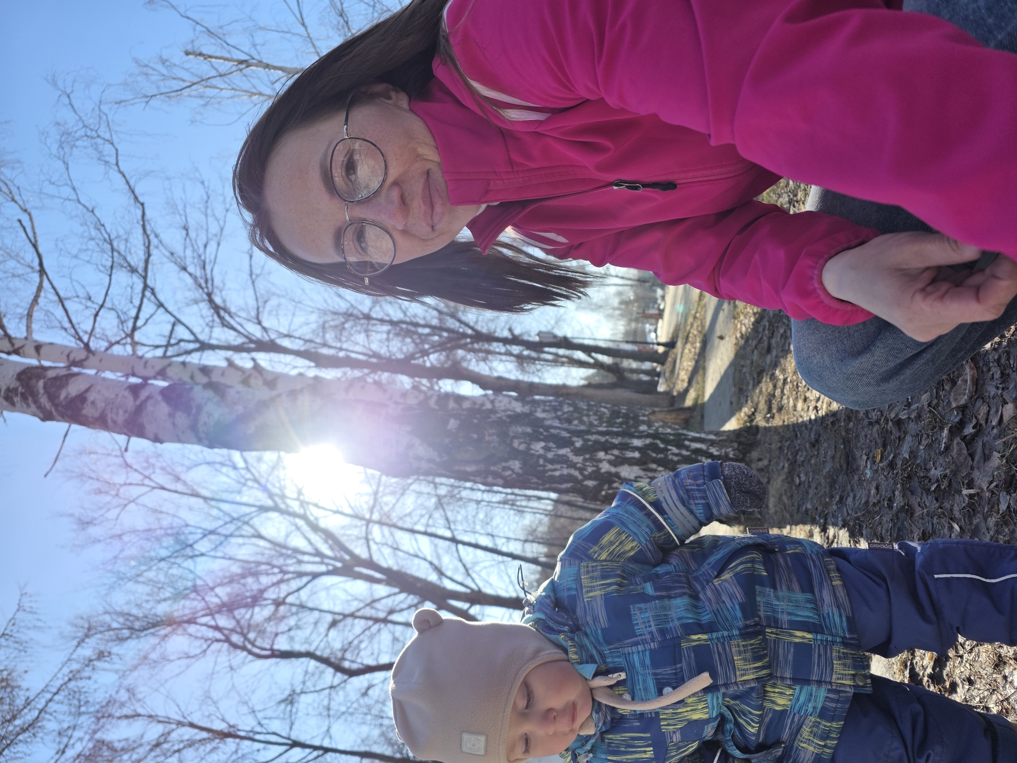 The image, taken in an outdoor setting in the city of Chaikovsky, Russia, shows a woman and a young child on what appears to be a partly sunny day.
The woman, wearing glasses and a bright pink jacket, is kneeling or squatting slightly above the child. Her dark hair is visible, and she seems to be looking down at the child. She is wearing jeans.
The child, who appears to be a toddler, is dressed in a blue and yellow plaid-patterned jacket and dark blue pants, along with a light brown hat with bear ears.
The ground is covered with dry leaves and grass. Several birch trees are visible in the background, with bare branches reaching towards a light blue sky. The sun is shining through the trees, creating a bright light with some glare. A narrow concrete path is seen in the middle background, running alongside the trees.