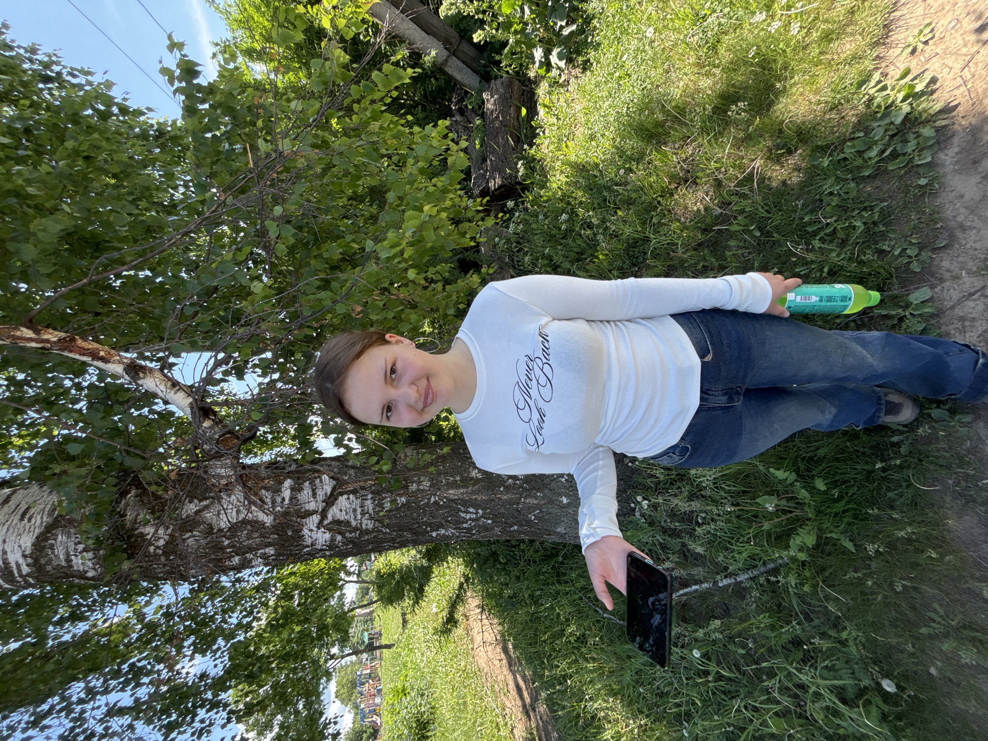 The image depicts a young adult female standing outdoors in a grassy area identified as Фоки, Russia. She possesses brown hair, a fair complexion, and a direct gaze at the viewer with a slight smile. Her attire consists of a white long-sleeved top bearing the black script text "Never Look Back" across the chest, blue denim jeans, and brown closed-toe footwear. In her outstretched left hand, she holds a dark-colored smartphone horizontally, its screen appearing off. Her right hand grasps a green plastic bottle. The environment is characterized by dense green grass and visible dirt paths. A prominent birch tree with distinctive white and dark bark stands immediately to her right. The background on the left reveals a distant children's playground with colorful structures, partially obscured by foliage. The right background features fallen logs and a weathered wooden post or beam among the undergrowth. The scene is illuminated by bright daylight under a clear blue sky, with faint power lines visible in the upper right quadrant.
