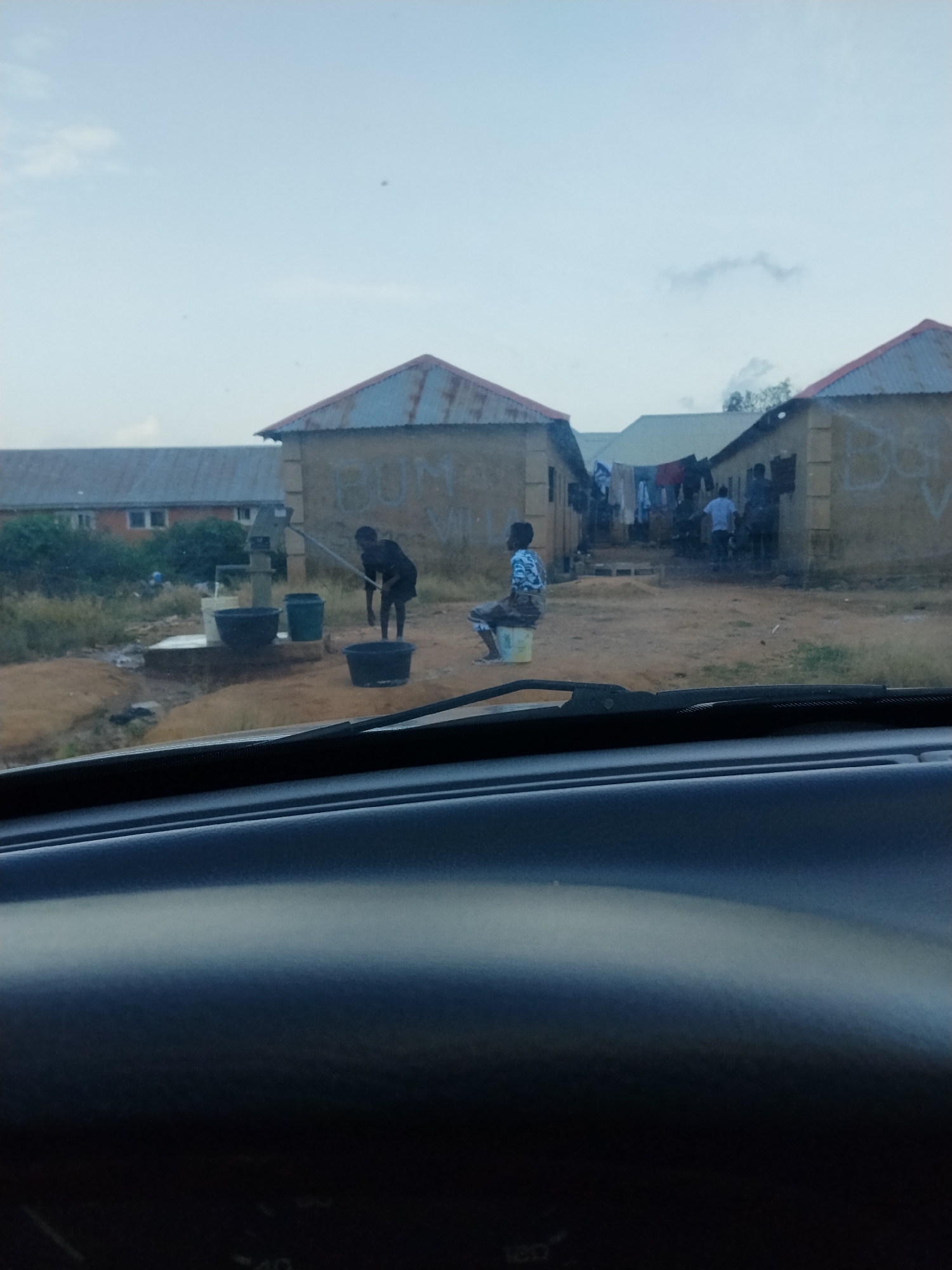 The image captures a scene in Durumi, Nigeria, likely a residential area. In the foreground, the dashboard of a car is visible, suggesting the photo was taken from inside a vehicle. The middle ground shows several single-story buildings with corrugated metal roofs. To the left, a water pump is in operation, with a person in a dark shirt and shorts drawing water into buckets. A person is also seen sitting on a bucket to the right of the water pump. Further into the scene, in front of one of the buildings, a person in a blue and white patterned shirt is seated on a yellow bucket. Clothes are hung out to dry on a line between buildings. The environment appears to be dusty, with sparse vegetation. The sky is overcast.